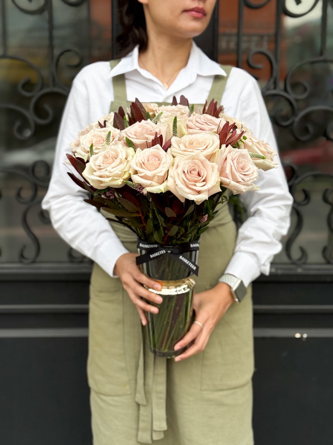 A person wearing a white shirt and olive green apron holds a bouquet of pale pink roses with dark foliage, evoking the charm of a Timeless Elegance Vase Arrangement, while standing in front of a decorative black metal gate.