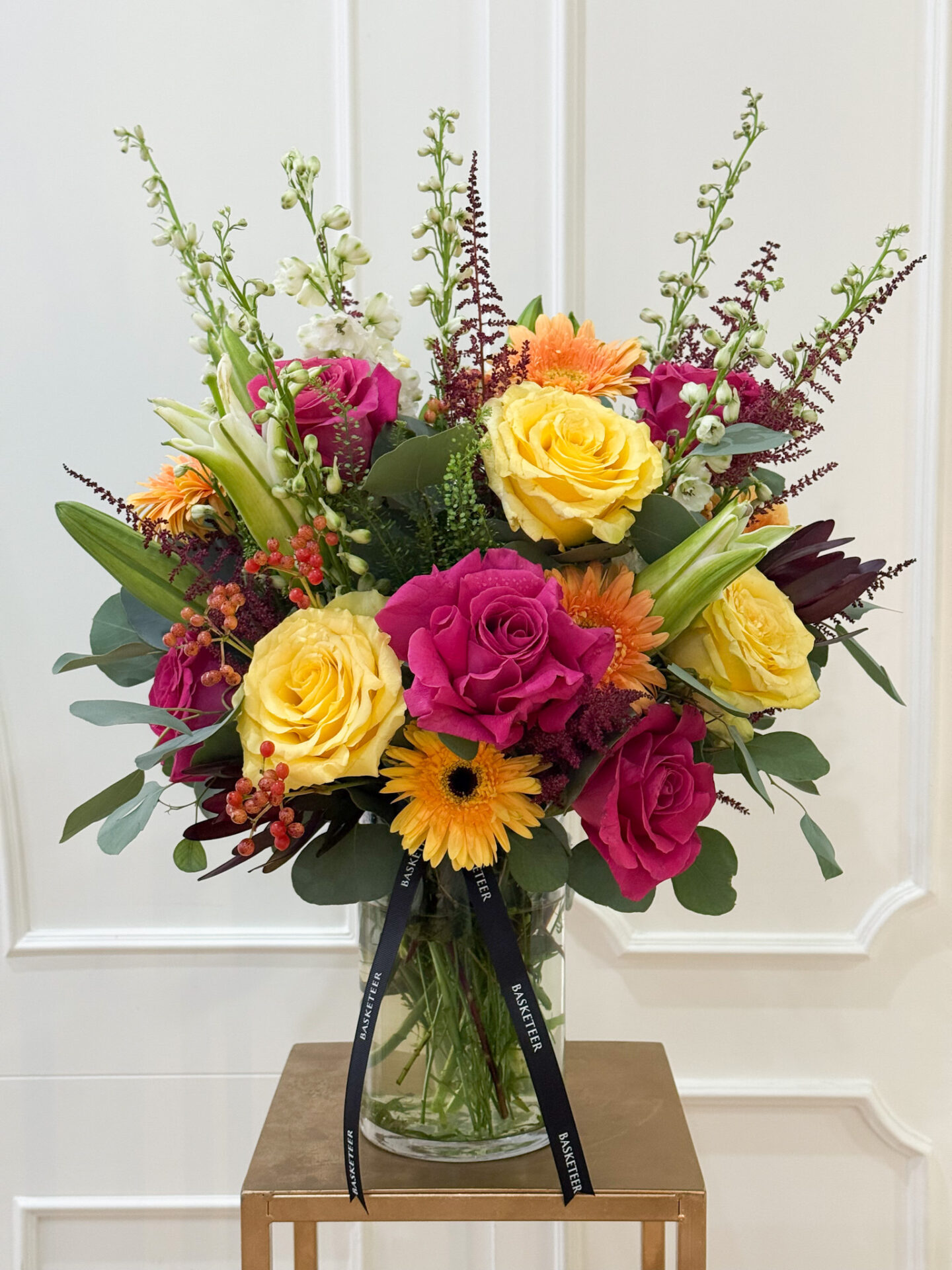 Bright floral vase arrangement with yellow roses, pink roses, orange gerberas, lilies, and greenery in a clear glass vase tied with Basketeer ribbon.