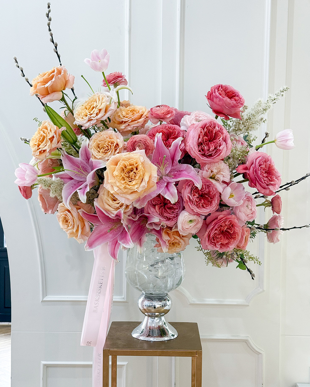 Elegant floral vase arrangement with pink lilies, peach roses, and pink garden roses in a tall glass vase with Basketeer ribbon.