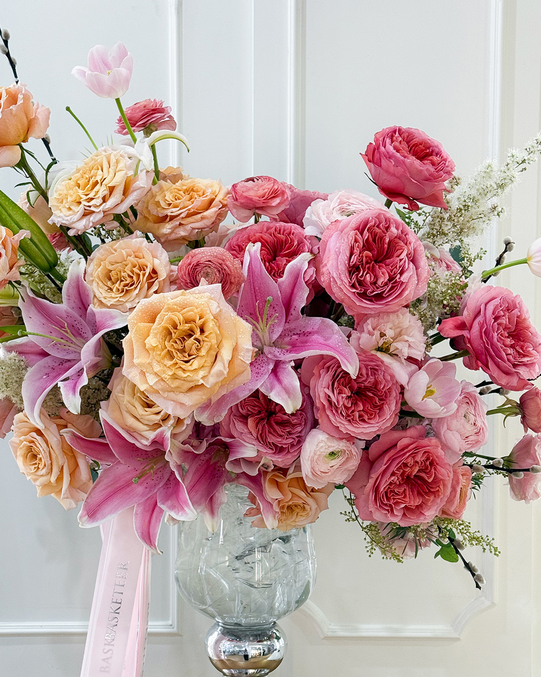 Elegant floral vase arrangement with pink lilies, peach roses, and pink garden roses in a tall glass vase with Basketeer ribbon.
