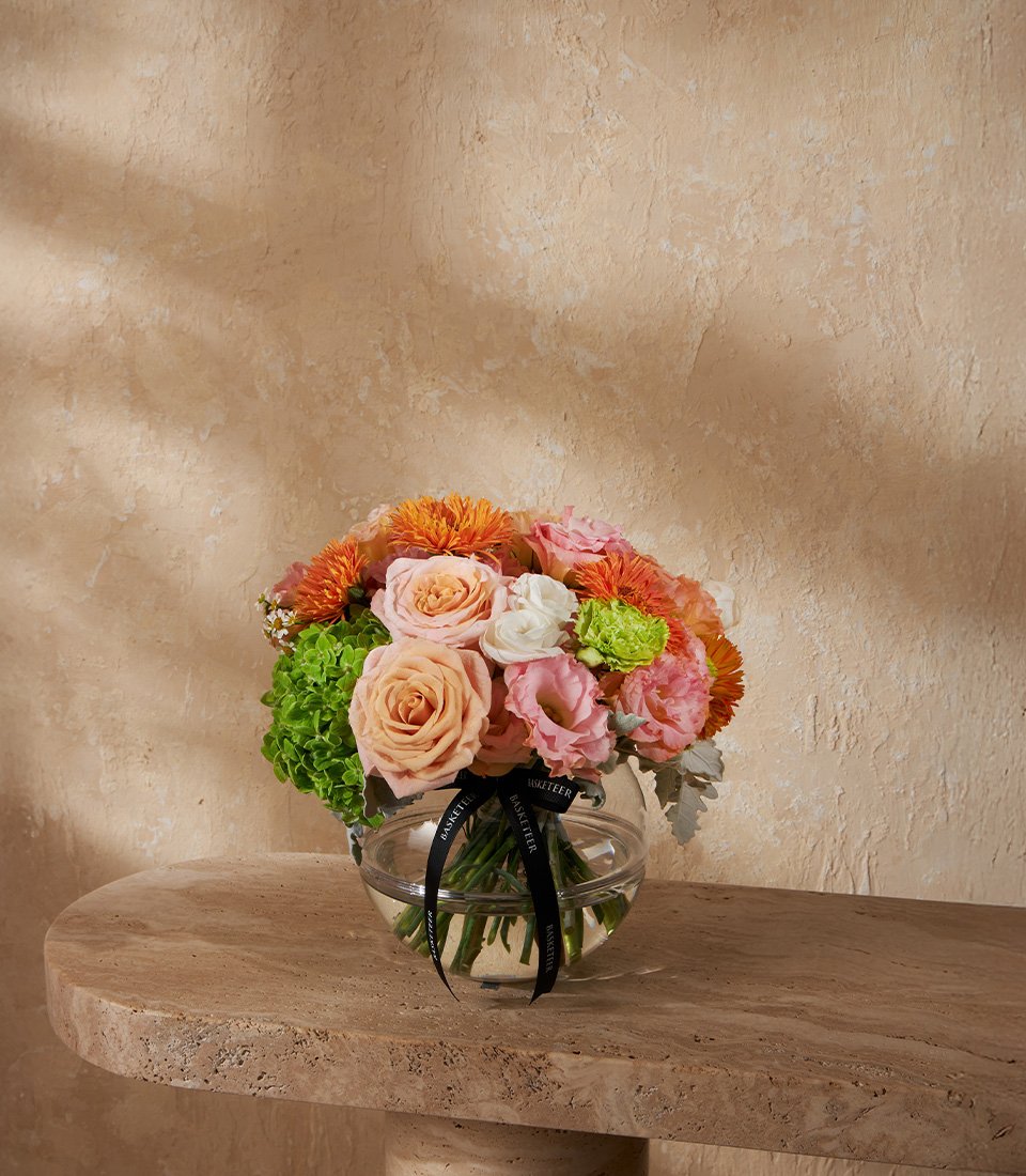 Charming flower arrangement with orange chrysanthemums peach roses green hydrangeas and pink lisianthus in a round glass vase Bangkok delivery