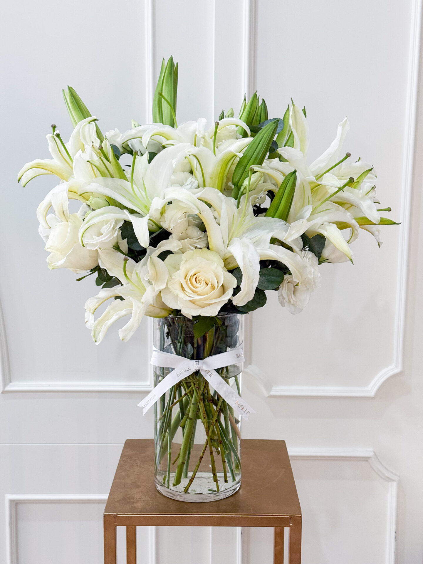 Elegant tall glass vase with white lilies and white roses, accented with greenery and tied with a white Basketeer ribbon.