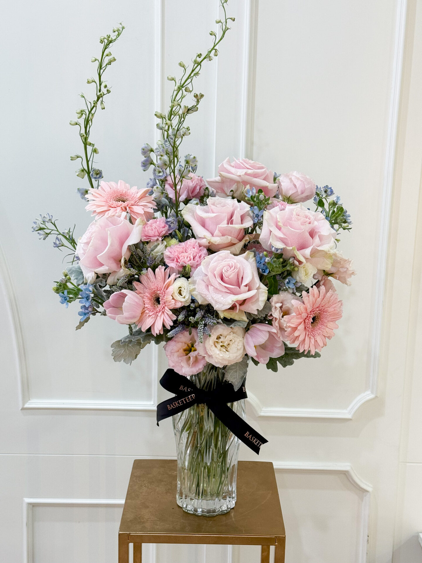 Elegant pastel flower vase arrangement with pink roses, pink gerberas, carnations, lisianthus, and soft blue flowers in a crystal vase with Basketeer ribbon.