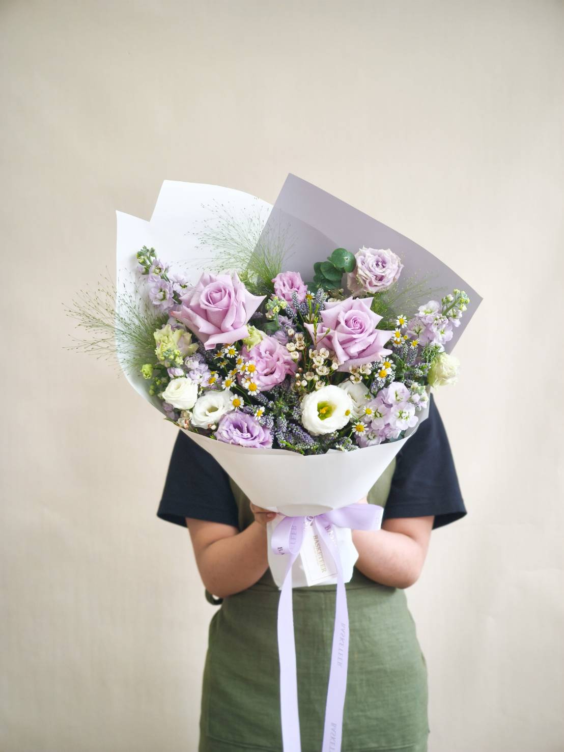 Elegant lavender bouquet with roses, lisianthus, white lisianthus, chamomile fillers, and greenery, wrapped in white paper and tied with lilac ribbon.