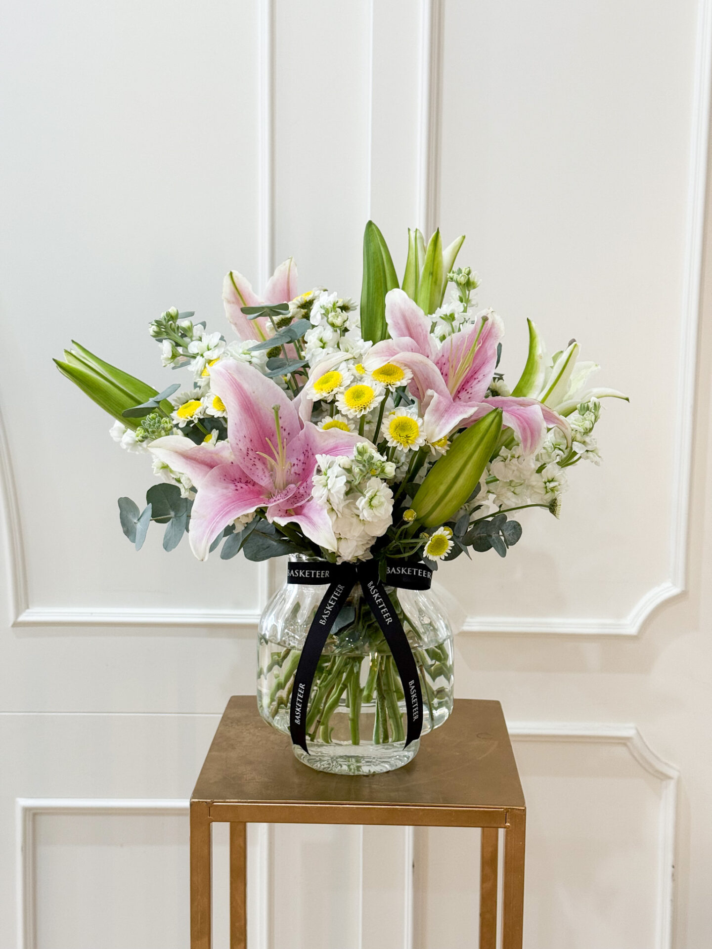 Elegant vase arrangement with pink lilies, white stock, chamomile, and eucalyptus greenery, styled in a clear glass vase with black satin ribbon.