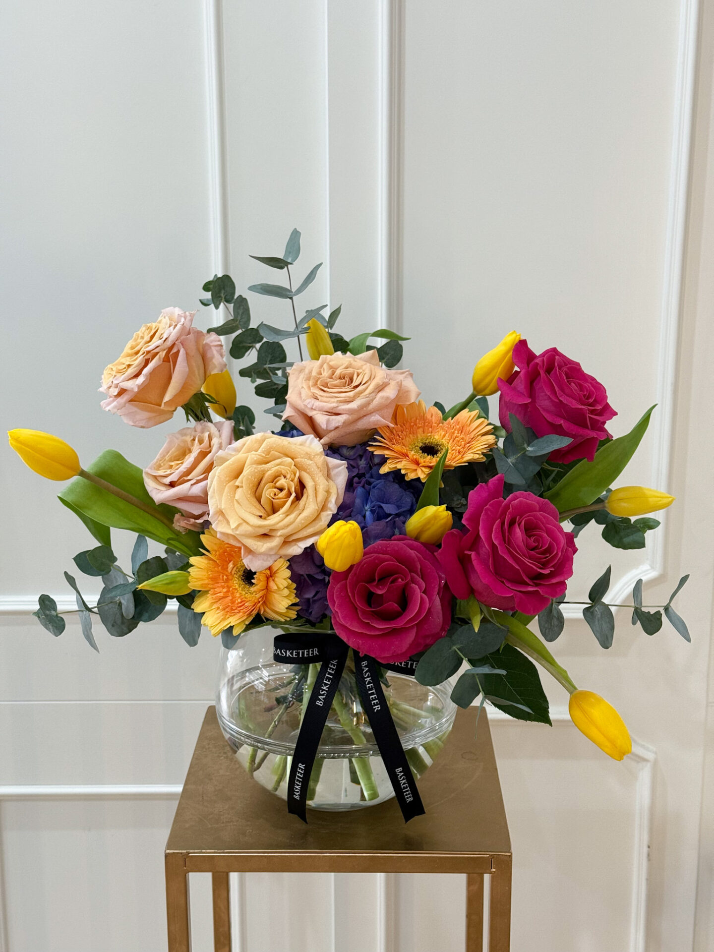 Elegant round glass vase with peach roses, fuchsia roses, yellow tulips, orange gerbera, blue hydrangea, and eucalyptus greenery tied with black satin ribbon.
