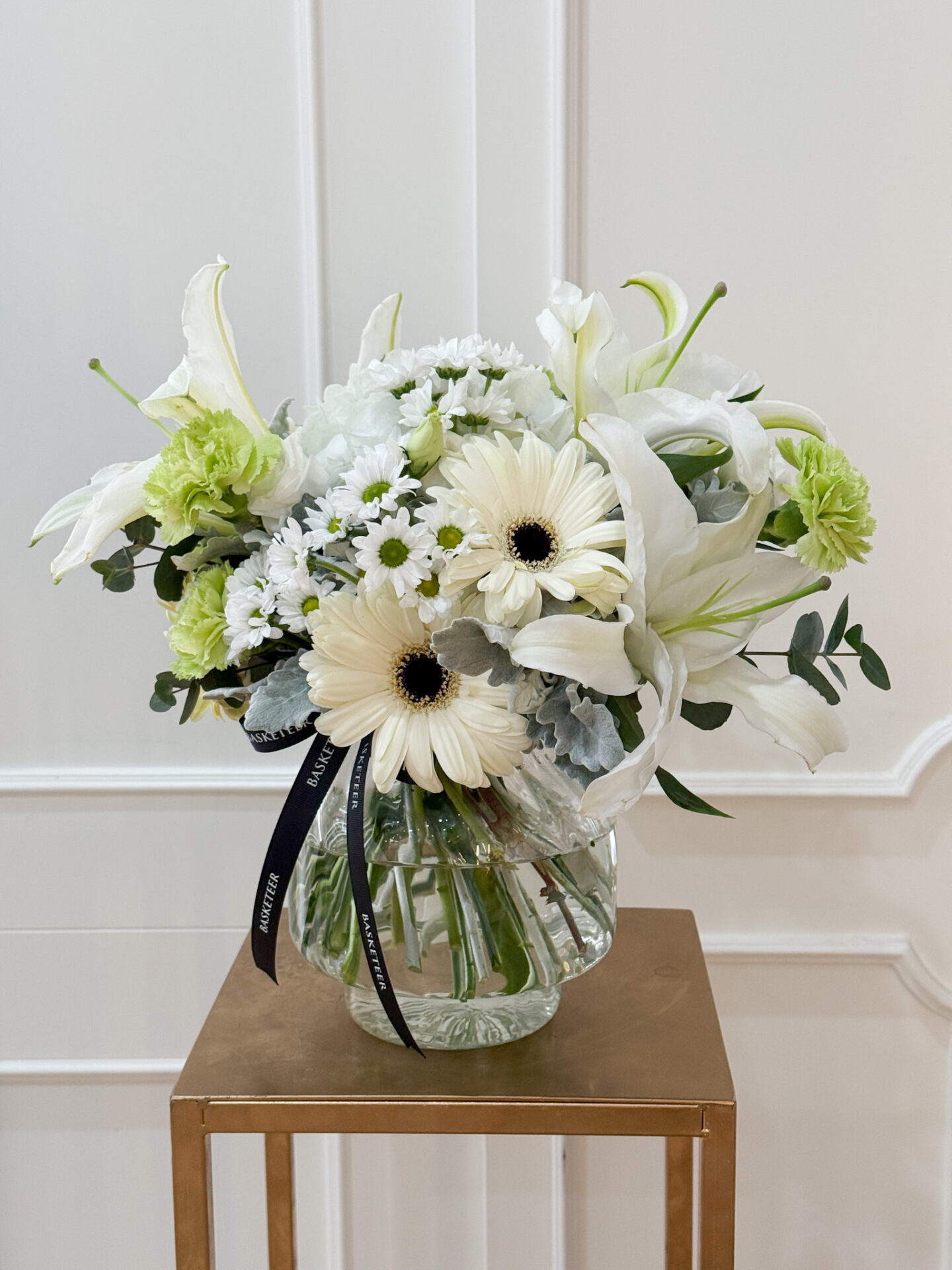 Elegant round glass vase with white lilies, white gerbera, green carnations, chamomile, and soft silver foliage, tied with a black satin ribbon.