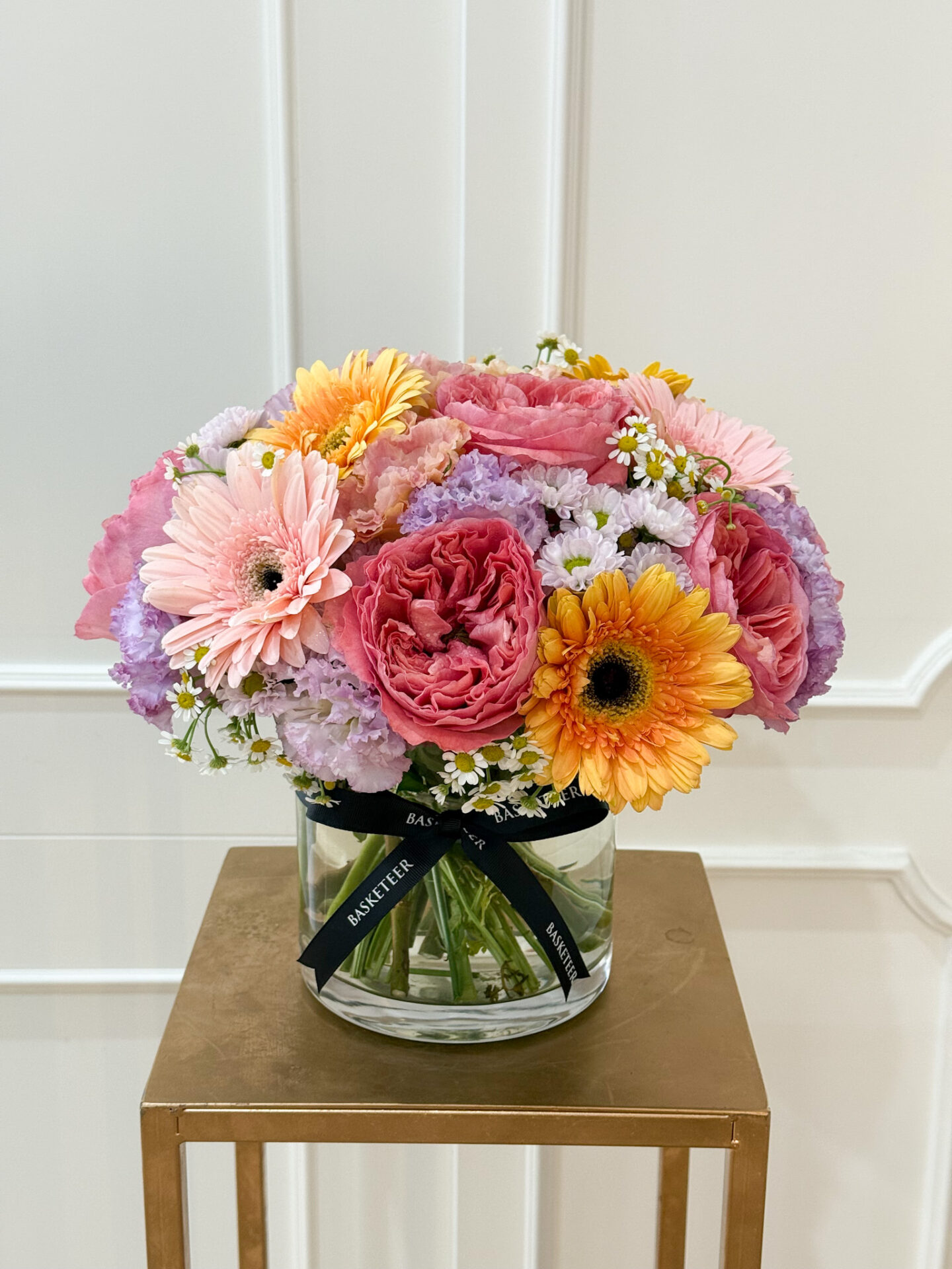 Vibrant glass vase with pink garden roses, orange gerbera, pastel pink gerbera, lavender statice, chamomile, and mixed seasonal blooms, tied with black satin ribbon.
