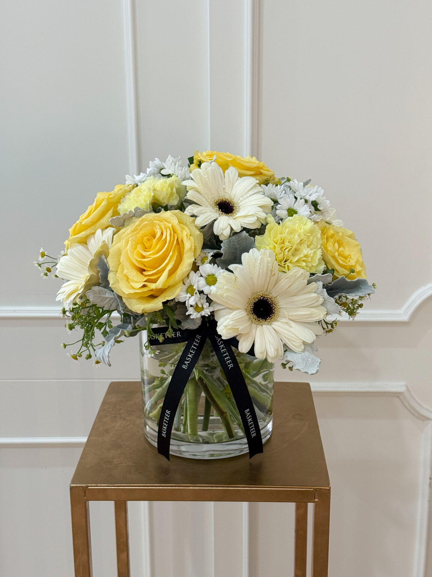 Elegant glass vase with yellow roses, white gerbera, yellow carnations, chamomile, and silver foliage, tied with a black satin ribbon.