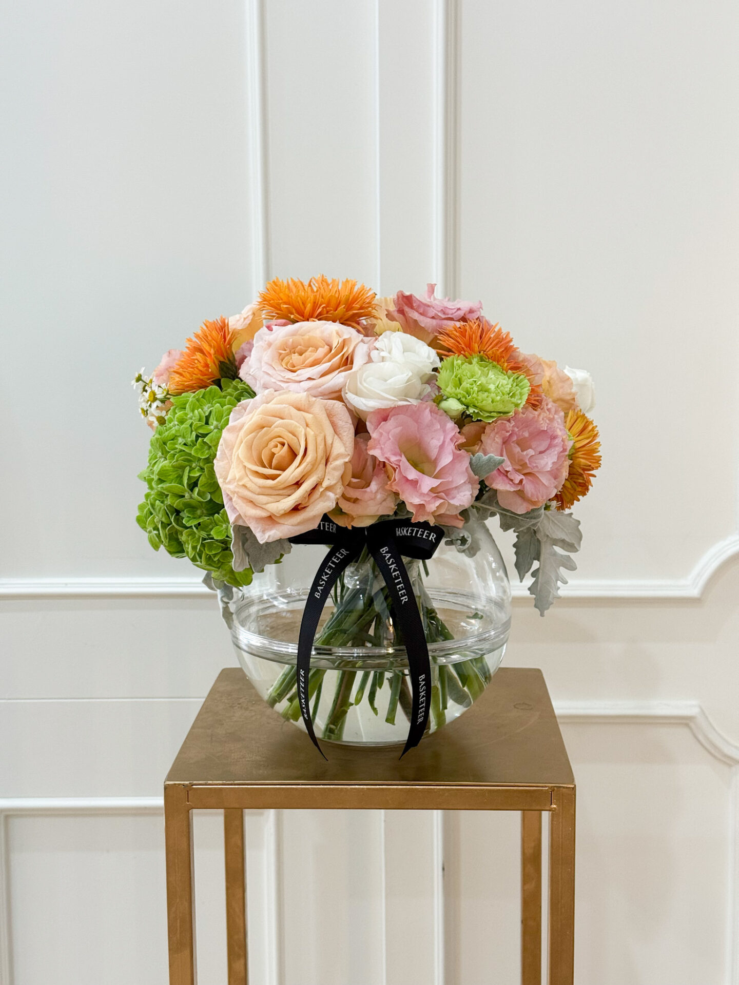 Elegant round glass vase with peach roses, green hydrangea, orange asters, pink lisianthus, and white ranunculus, tied with black satin ribbon.