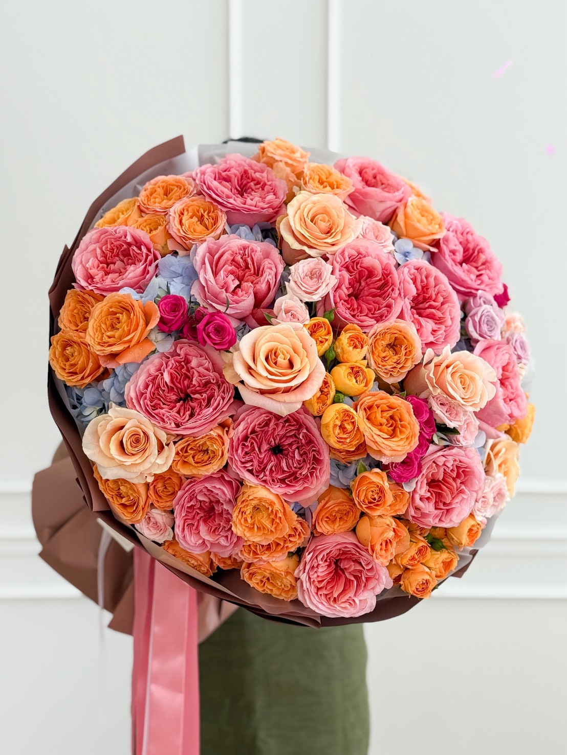The Luxury Harmony Rose Bouquet features a large arrangement of roses in pink, peach, orange, and light purple, wrapped in brown paper and tied with a pink ribbon, held against a white background.