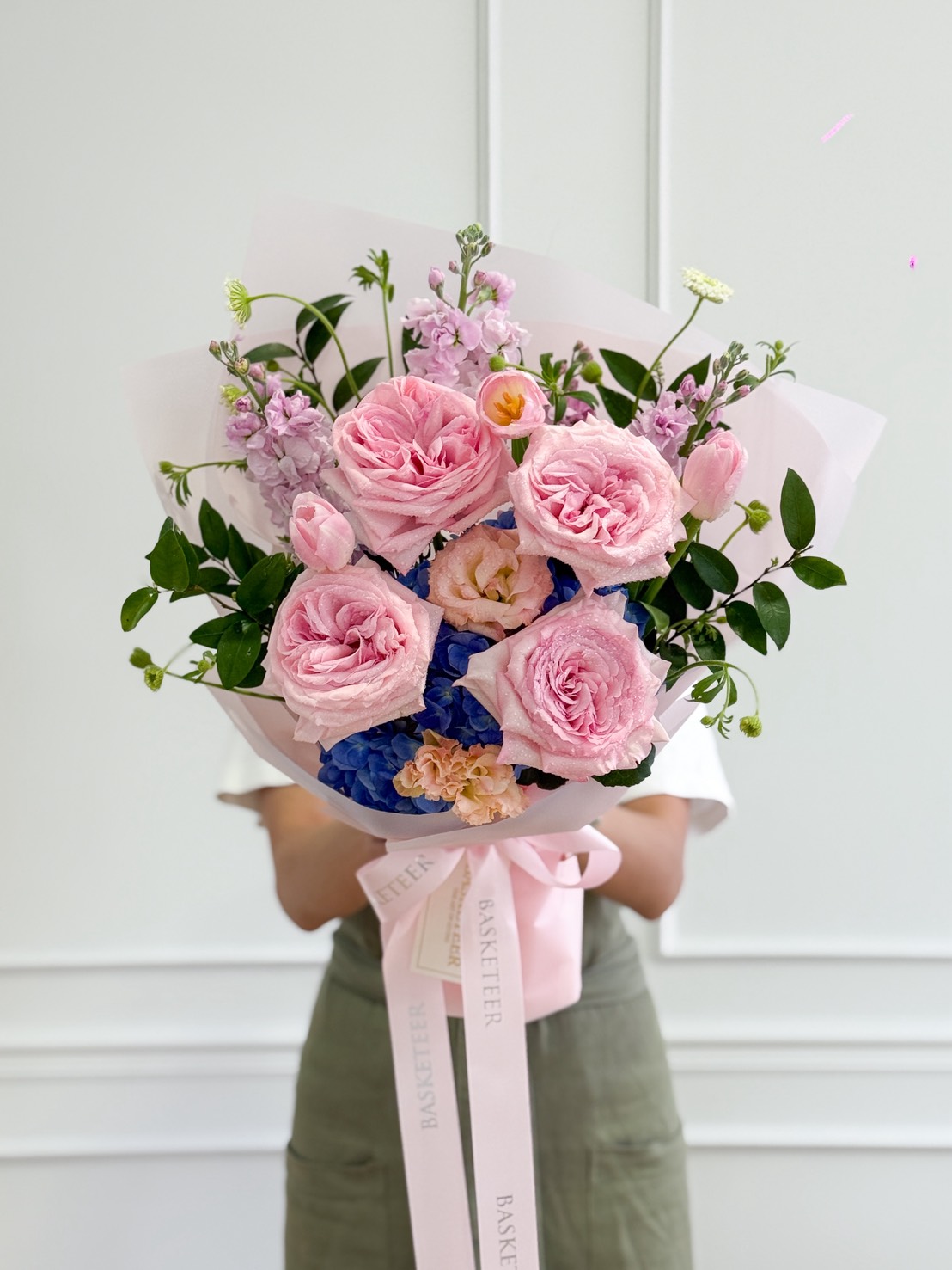 A person in a light shirt and green apron holds the Blush Serenity Flower Bouquet—pink roses, blue hydrangeas, and assorted greenery—wrapped in pale pink paper with a “BASKETEER” ribbon, set against a white wall.