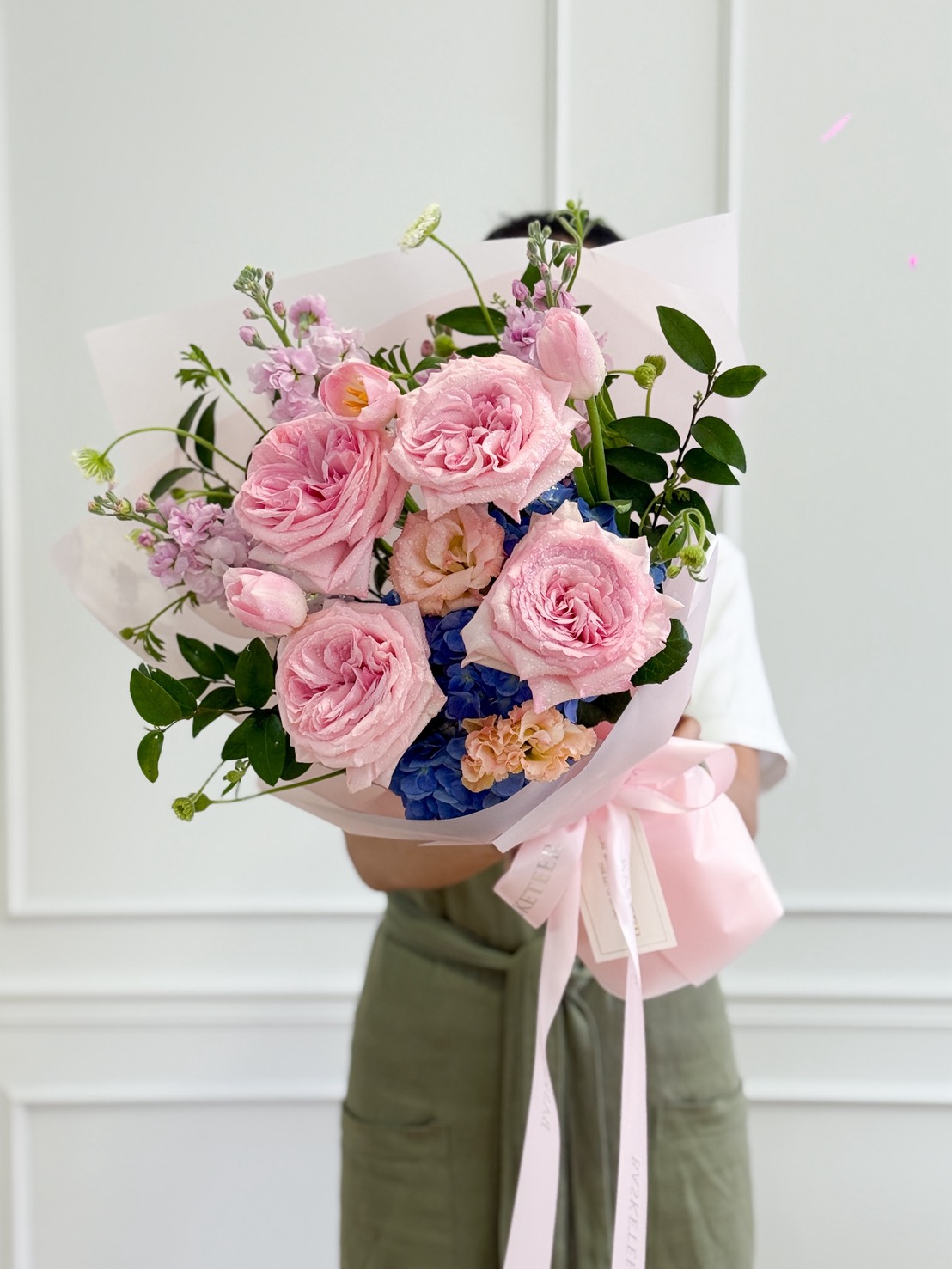 A person wearing a white shirt and green apron holds the Blush Serenity Flower Bouquet—pink roses, light pink tulips, purple flowers, and greenery wrapped in pale paper with a pink ribbon—against a white wall background.