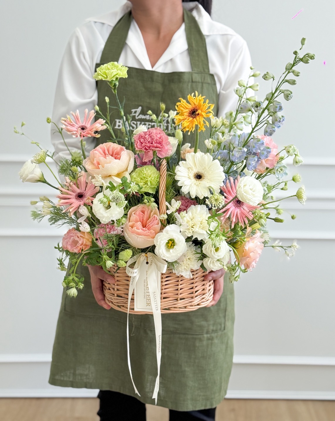 Large pastel flower basket with roses, gerbera, ranunculus, carnations, lisianthus, delphinium, and mixed blooms, held by florist in green apron