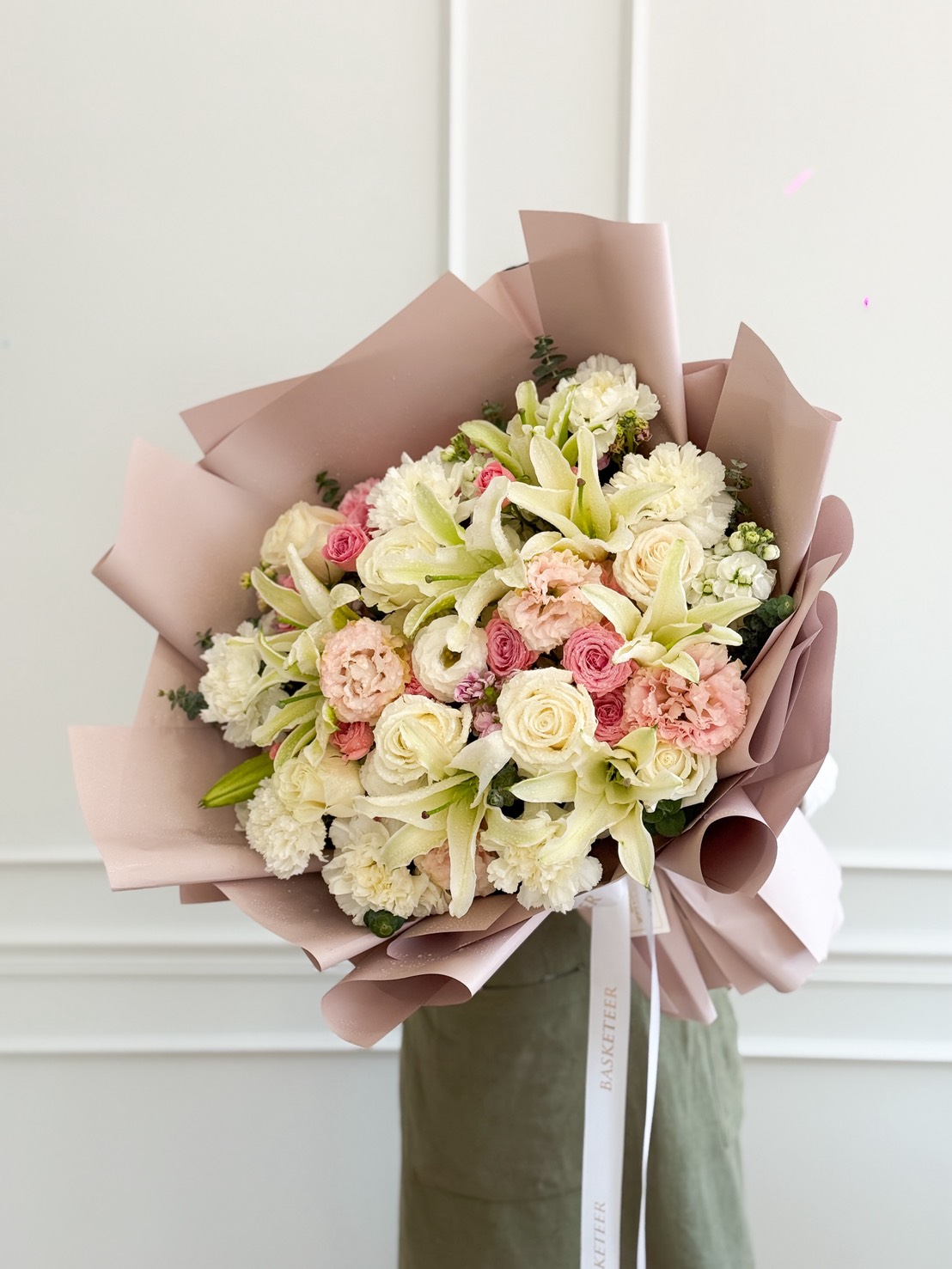 A large Ivory & Blush Roses with White Tulips Pastel Bouquet, featuring white lilies, pink and white roses, and other blooms wrapped in light pink paper, held by a person in a green outfit against a white wall.