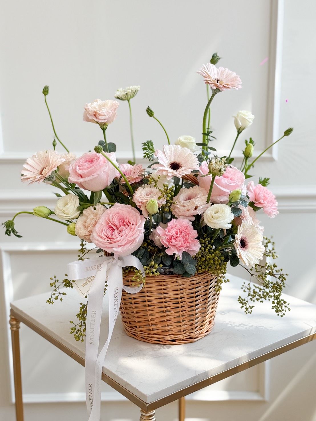 The Blush Harmony Flower Basket, filled with pink and white roses and gerbera daisies, sits on a small white table. A white ribbon with text is tied round the handle, whilst the softly lit background adds a gentle touch.