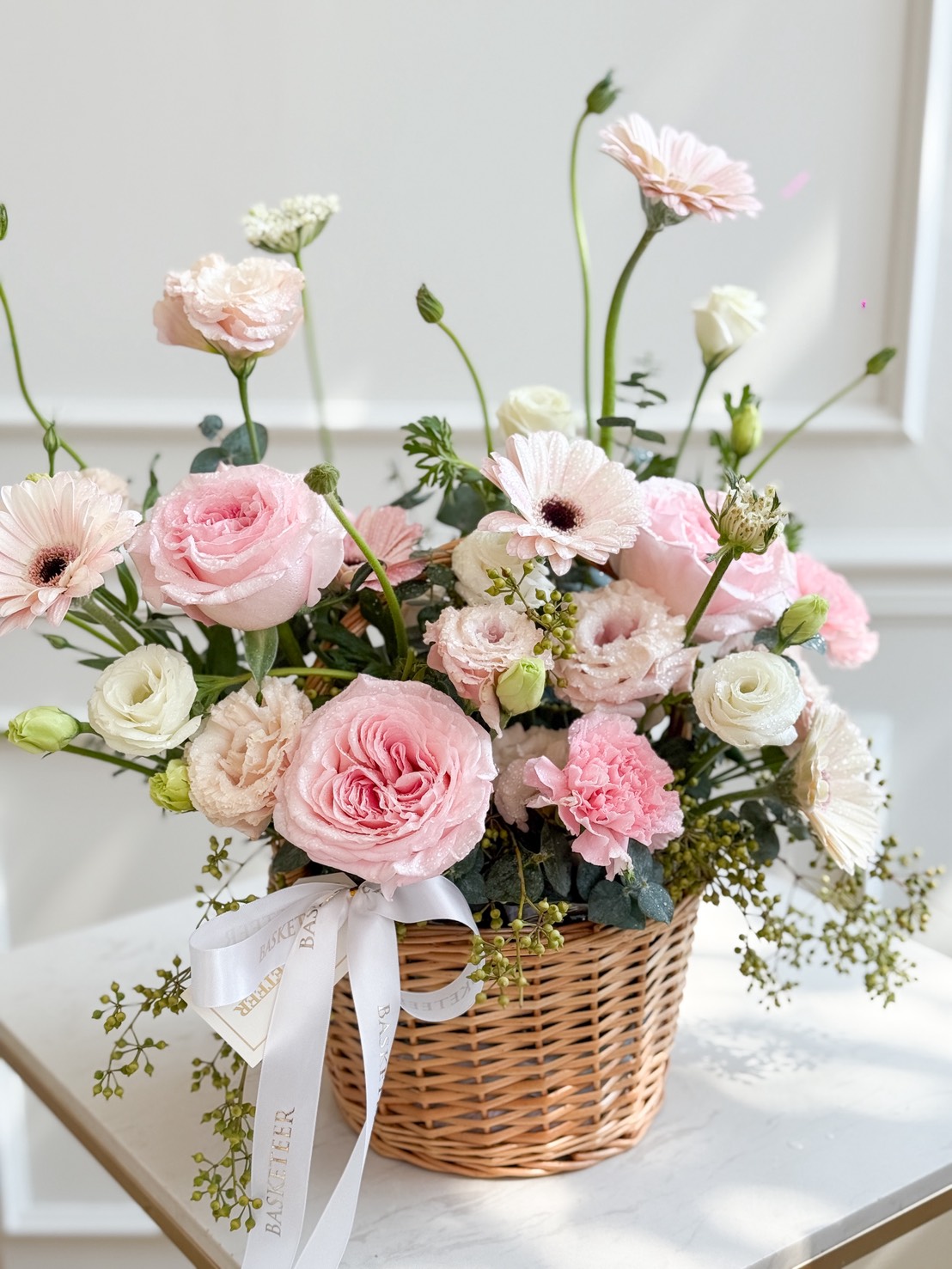 The Blush Harmony Flower Basket features a wicker basket filled with pink and white roses, gerbera daisies, and lush greenery, all accented by a delicate white ribbon. This elegant arrangement rests beautifully on a crisp white surface.