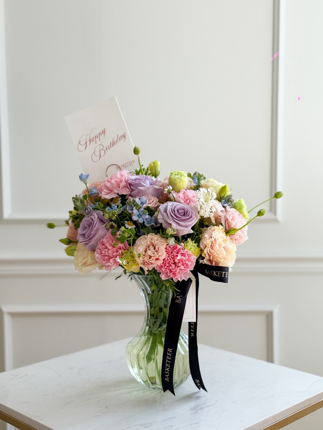 A Blush Harmony Vase filled with pastel flowers—pink, purple, and cream blooms—sits on a white table. A 