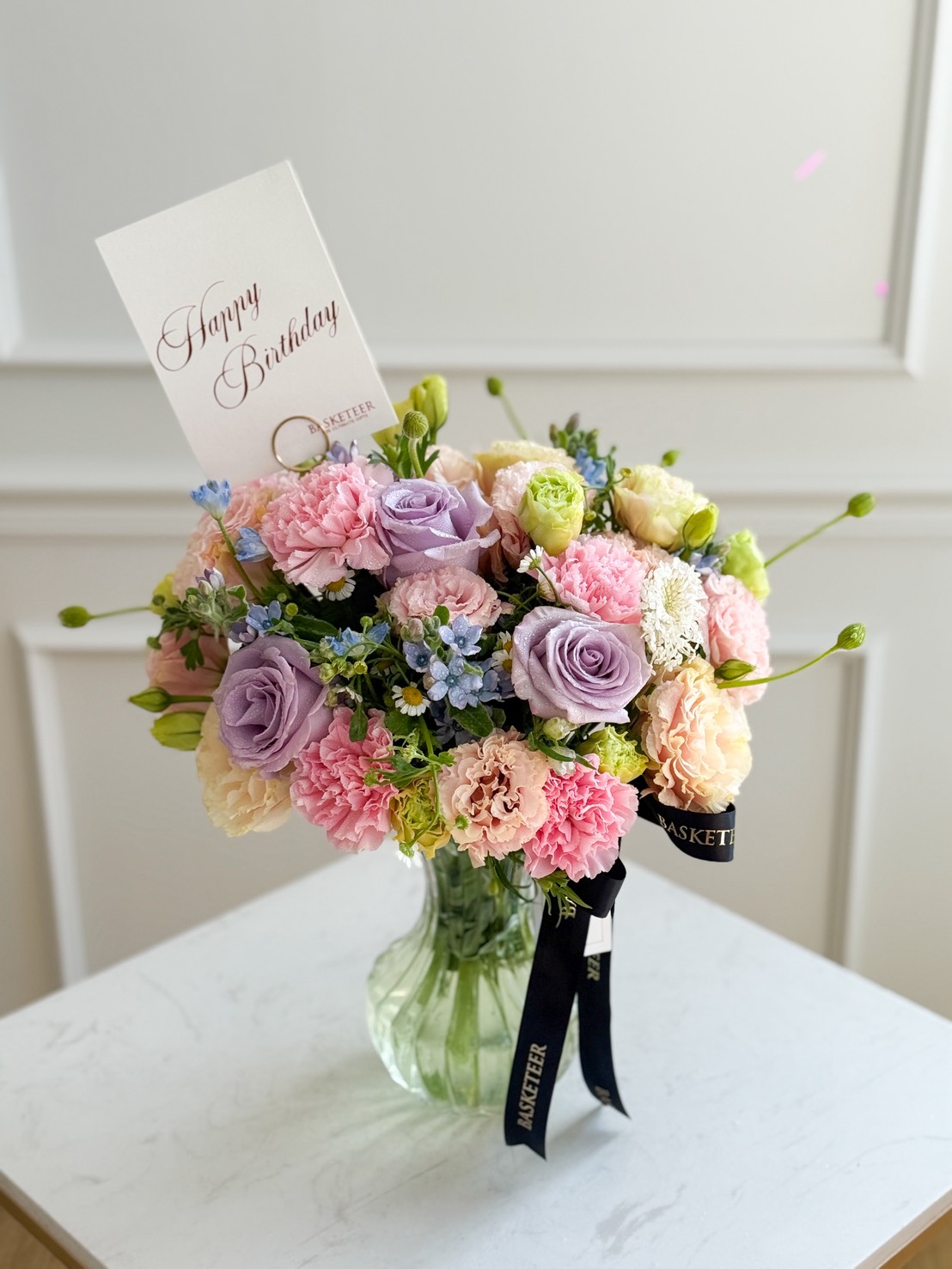 A Blush Harmony Vase holds a bouquet of pastel flowers, including pink carnations and purple roses, with a “Happy Birthday” card tucked in. The vase is decorated with a black ribbon and sits elegantly on a white surface.
