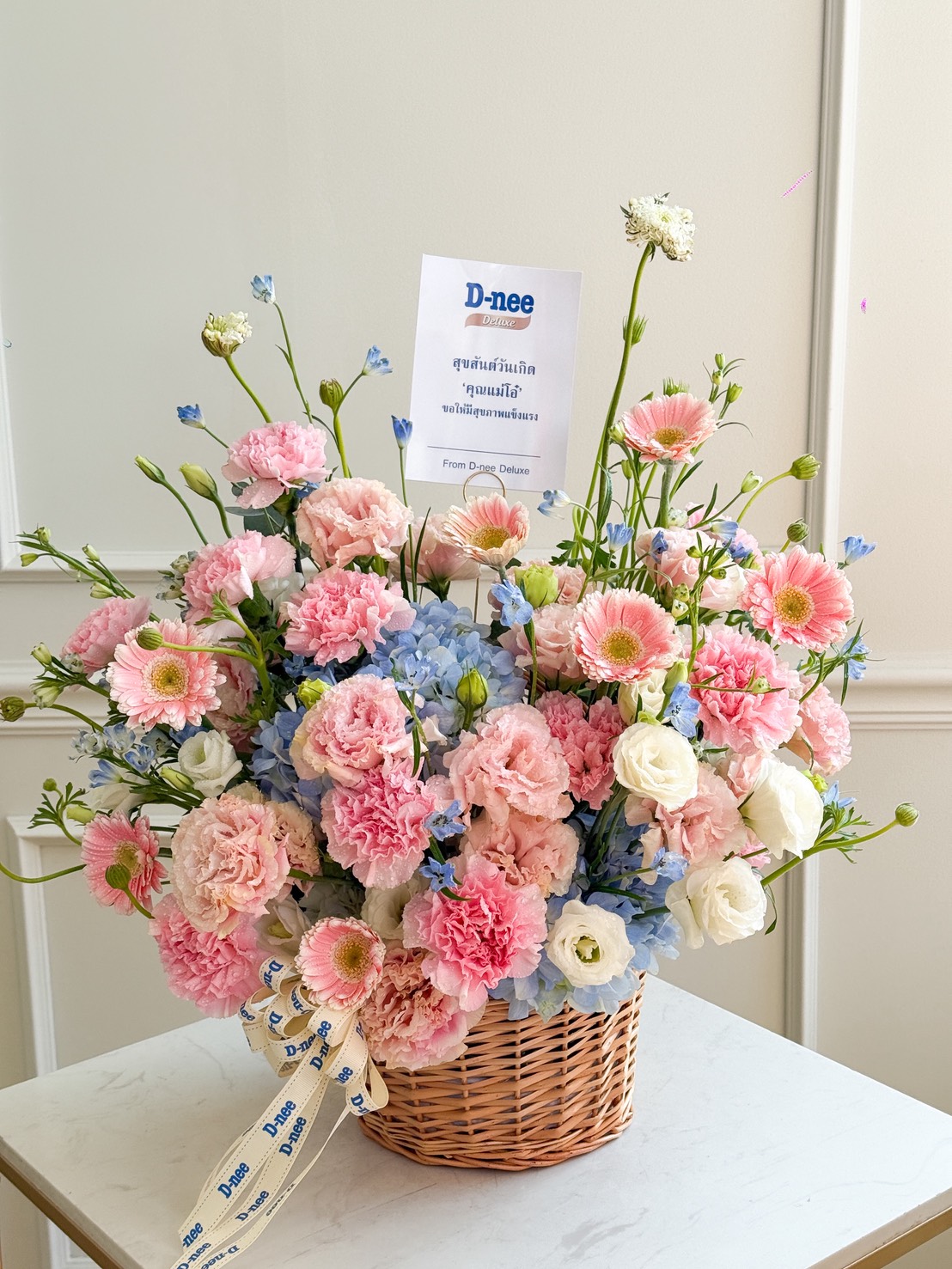 The Pastel Radiance Flower Basket features a wicker basket filled with pink carnations, white roses, blue hydrangeas, and pink gerbera daisies. A 