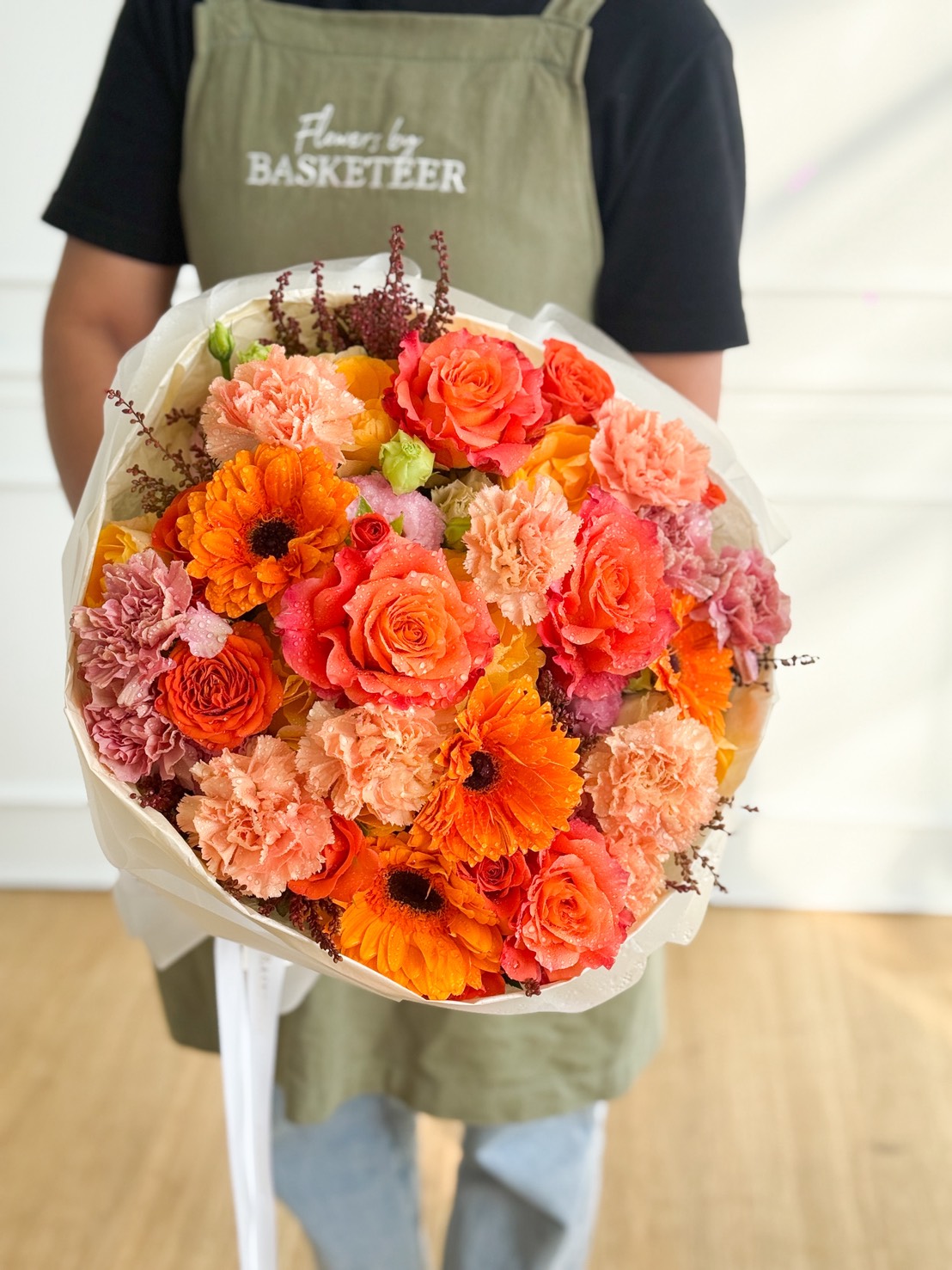 A person in a green apron holds the Radiant Flame Mixed Bouquet—vibrant orange and peach roses, carnations, and gerbera daisies—wrapped in white paper. The softly lit, minimalistic background highlights the bouquet’s beauty.