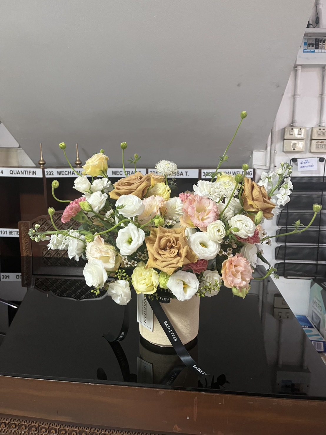 A Golden Toffee Elegance Vase holds a floral arrangement of yellow, white, and peach roses with green stems and small white flowers, set on a black glossy surface with shelves and labels in the background.