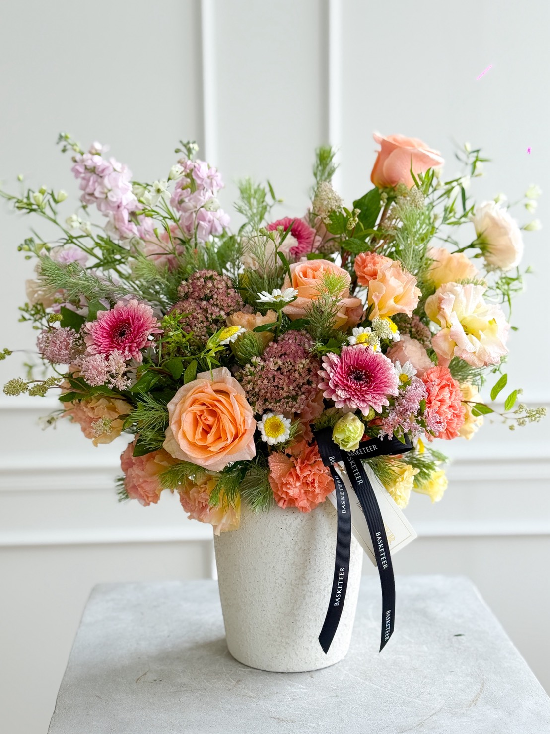 A white Pastel Garden Symphony Vase holds a colourful bouquet of roses, chrysanthemums, carnations, and assorted greenery, accented with a black ribbon that reads 