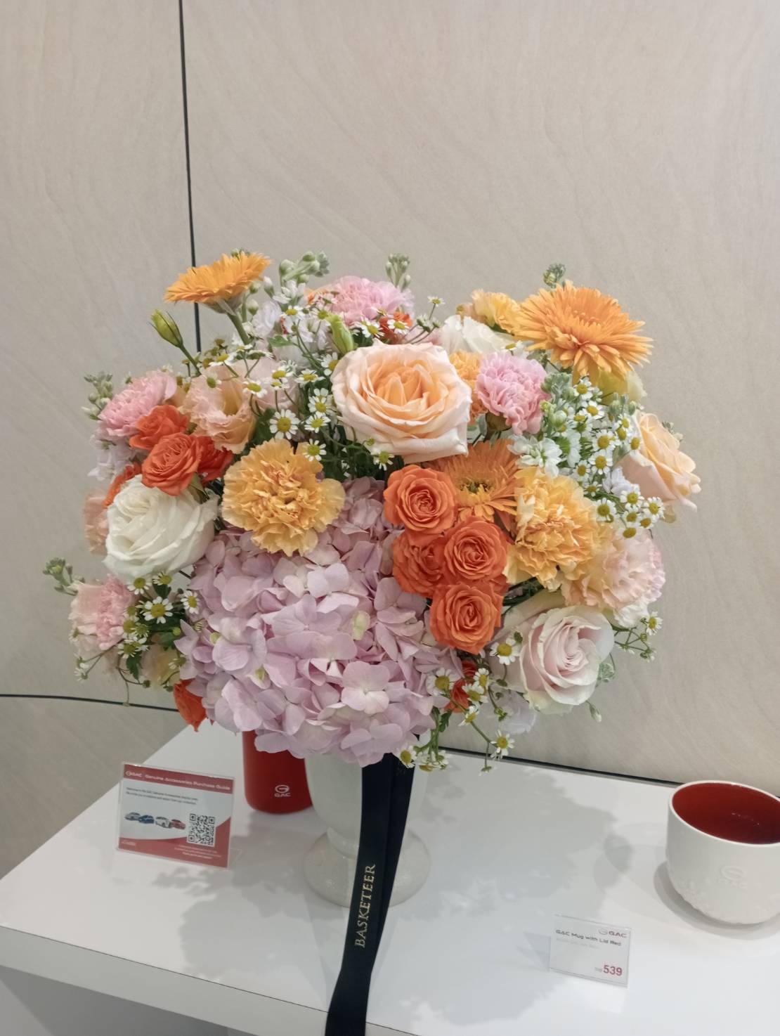 The Golden Dawn Vase Arrangement features a bouquet of pastel flowers—roses, carnations, and hydrangeas in pink, orange, yellow, and white—arranged in a white vase on a white table beside a red cup and small price tags.