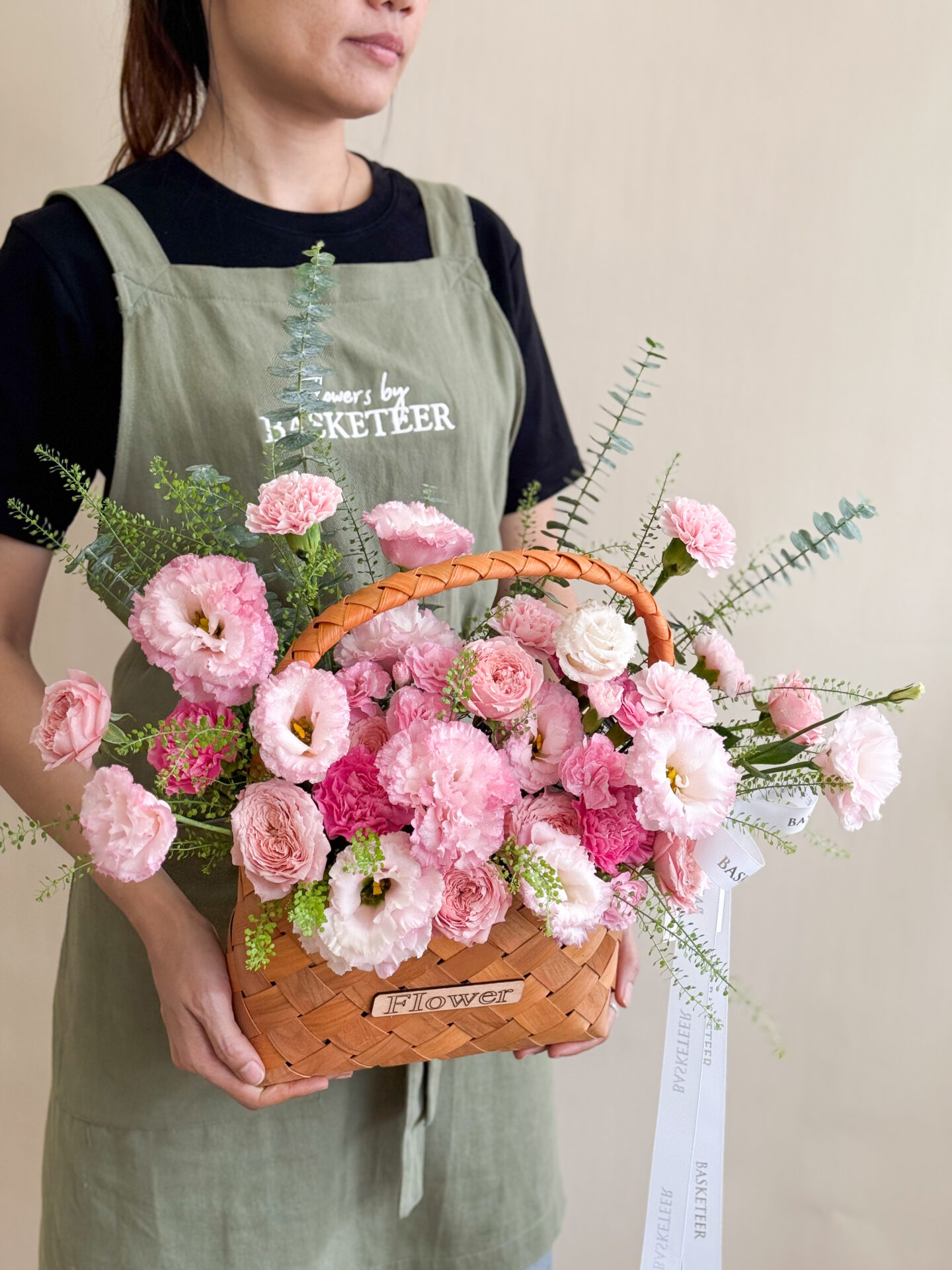 A person in a green apron holds the Pink Delight Flower Basket, filled with pink roses, lisianthus, and greenery. The basket features a 