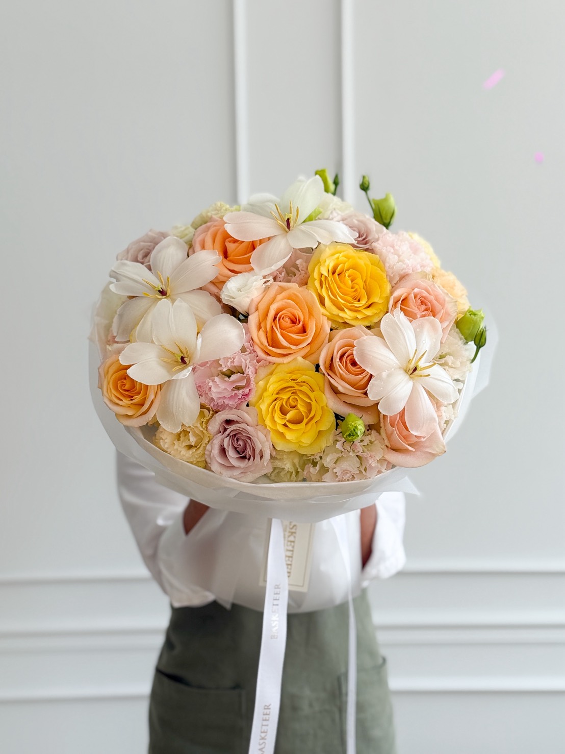 A person holding a Serene Grace Mixed Bouquet featuring yellow and peach roses, white lilies, and pale pink blooms, set against a white background with decorative moulding.