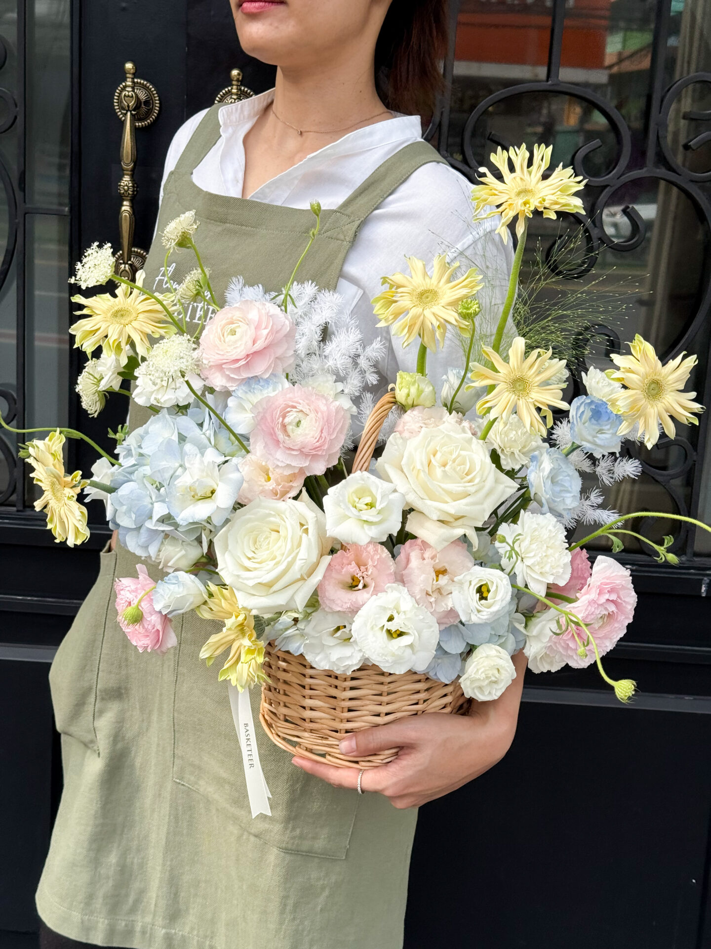 A person in a light green apron and white shirt holds a Pastel Radiance Flower Basket filled with white roses and yellow daisies, standing against a black decorative door.