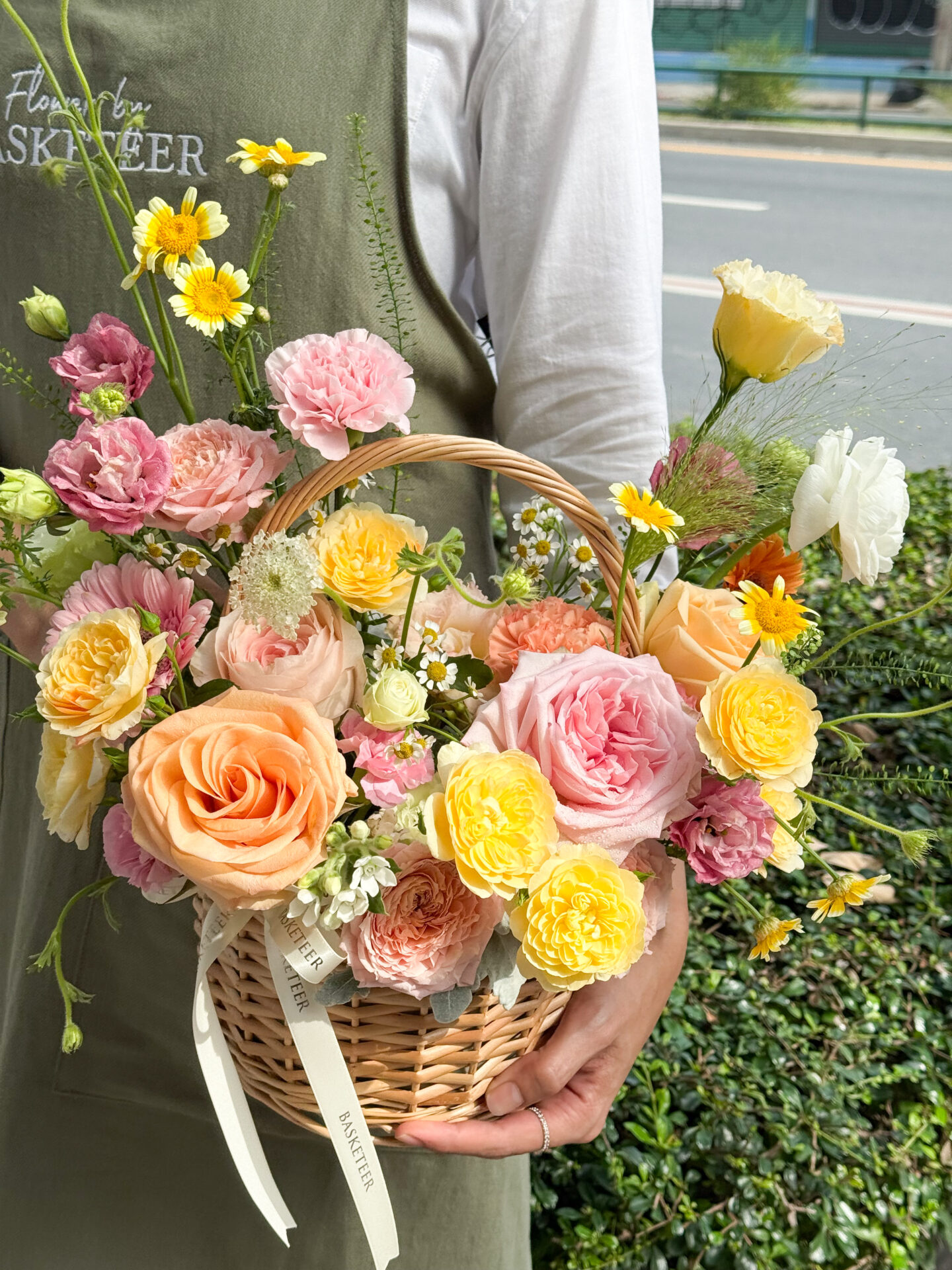 A person in a green apron holds the Radiant Harmony Flower Basket filled with assorted fresh flowers—pink, yellow, and white roses and carnations—against a leafy outdoor backdrop.