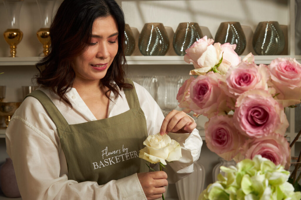 A florist wearing an apron with the Flowers by Basketeer logo, arranging white and pink roses.