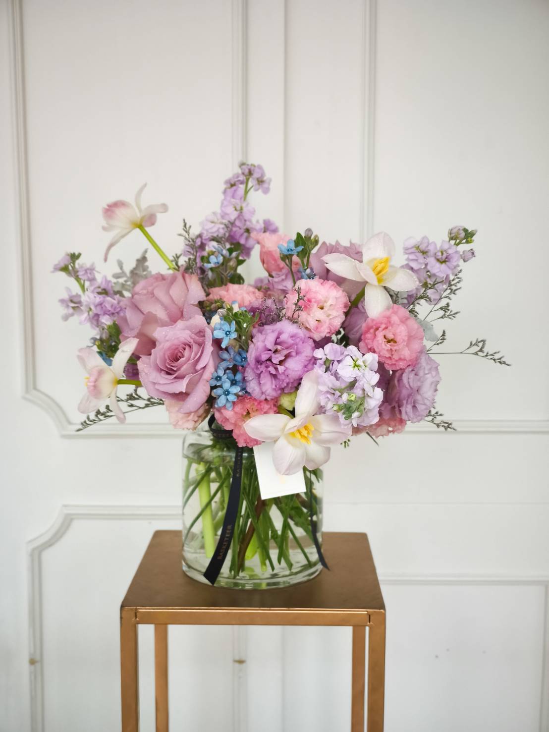 Elegant glass vase with pastel purple roses, pink carnations, white daffodils, and soft blue accents.
