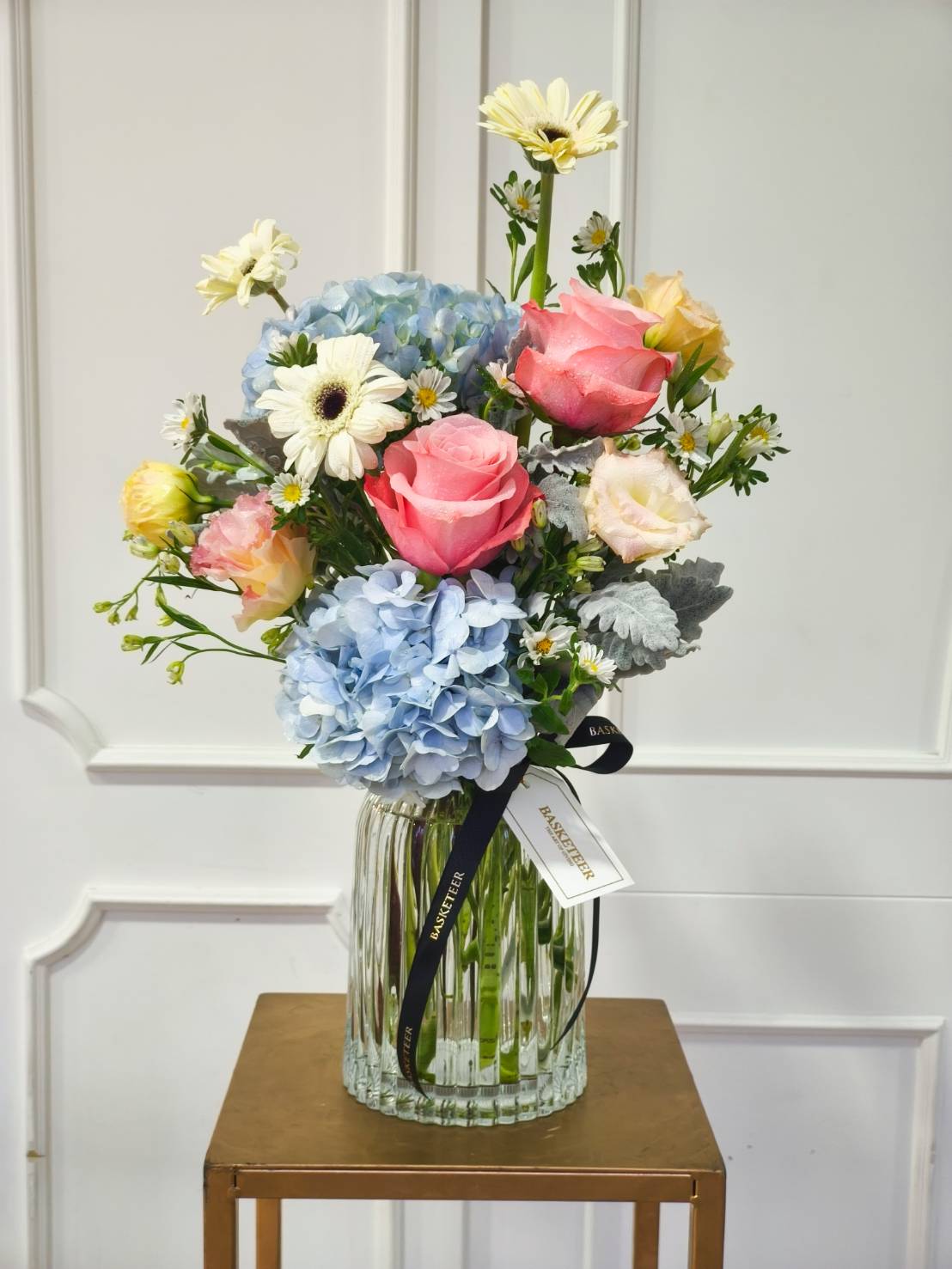 Elegant glass vase with pink roses, blue hydrangeas, white gerberas, and pastel seasonal blooms.