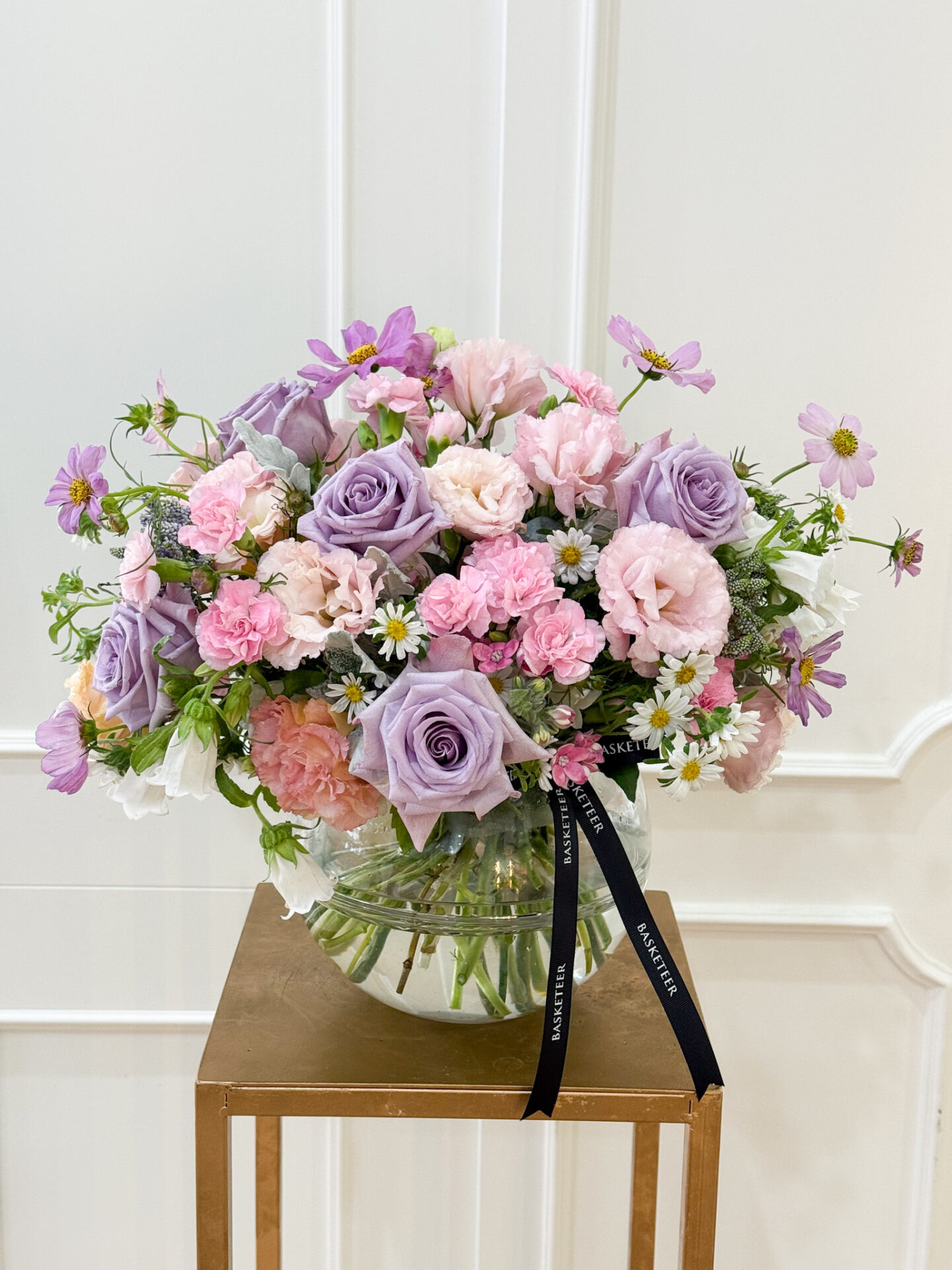 Glass vase with lavender roses, pink carnations, lisianthus, cosmos, daisies, and pastel blooms arranged elegantly.