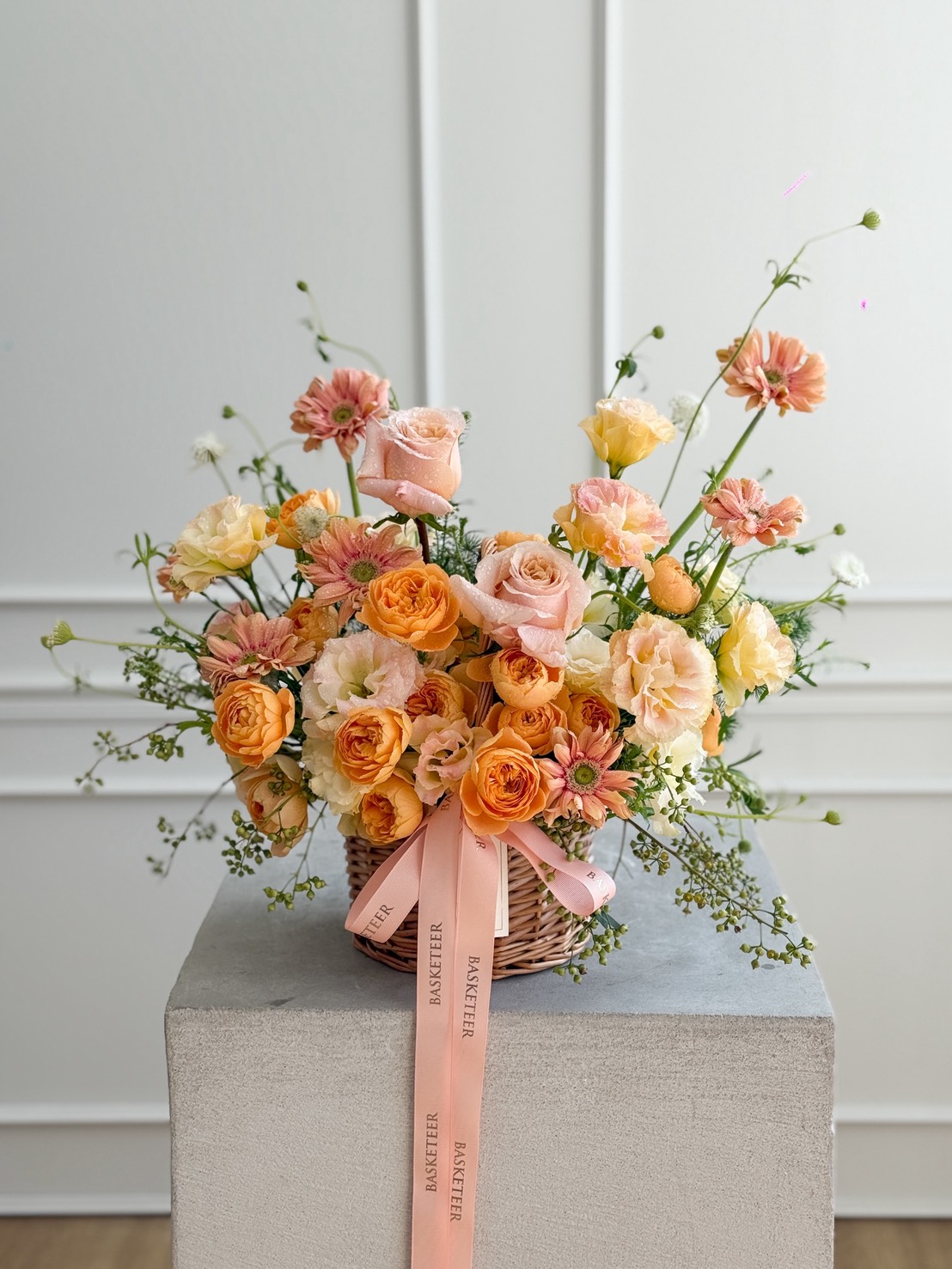 A basket of peach, orange, and pale yellow roses and flowers, accented with greenery, sits on a grey pedestal. This Sunset Glow Flower Basket is adorned with a pink ribbon that reads 