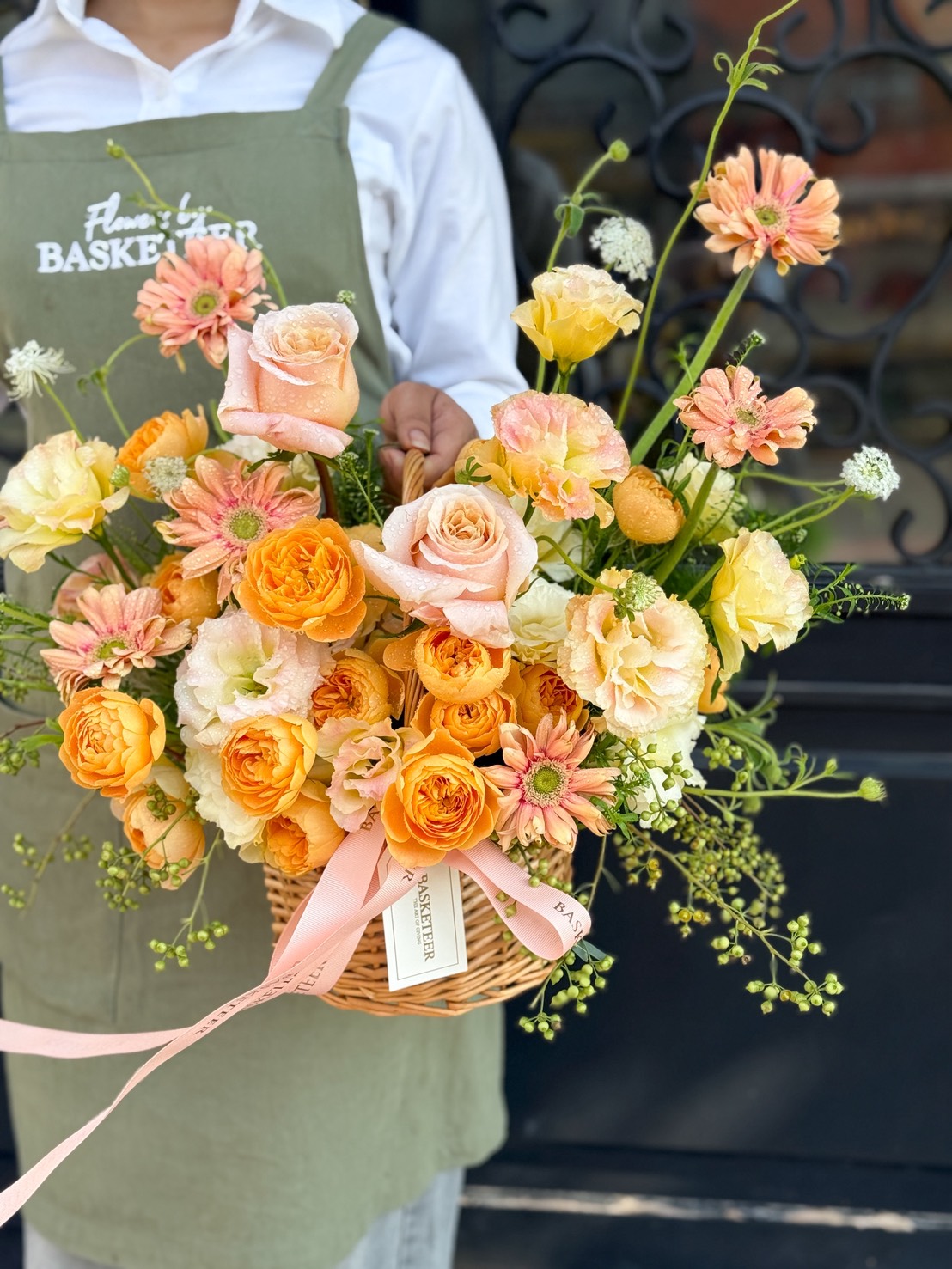 A person wearing a green apron holds the Sunset Glow Flower Basket—filled with orange, yellow, and pink flowers accented with greenery and pink ribbon—standing in front of a black ornate door.