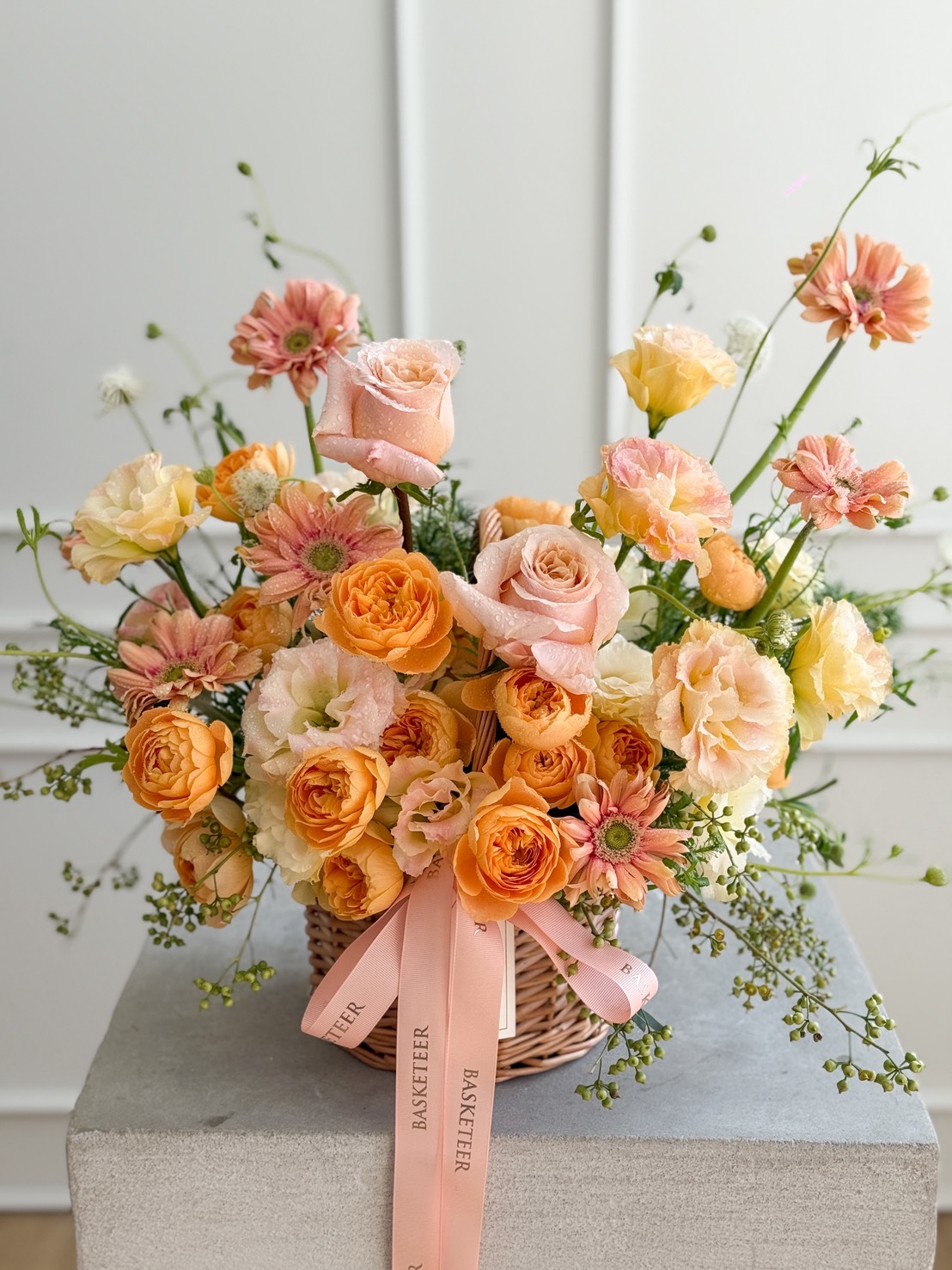 The Sunset Glow Flower Basket features a woven basket filled with peach, orange, and pale pink blooms, accented with greenery and tied with a pale pink ribbon, sitting gracefully on a grey surface against a white background.