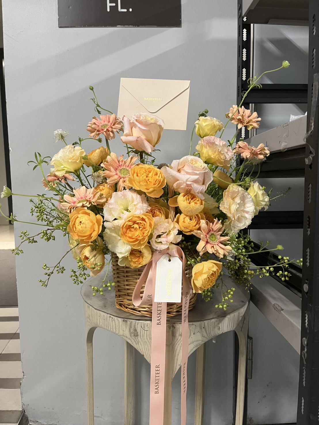 A wicker basket filled with a vibrant Sunset Glow Flower Basket—yellow, peach, and pink blooms—sits on a small table. A cream envelope is tucked among the flowers, with a pink ribbon and tag hanging from the basket.