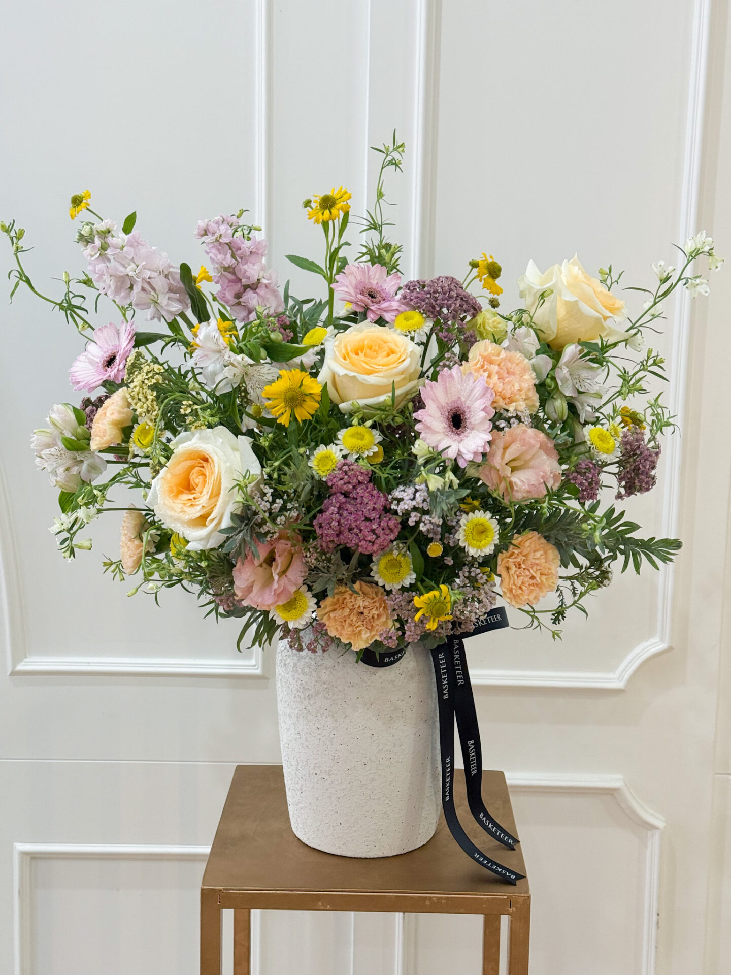 Cheerful garden-style vase with yellow roses, pastel gerberas, carnations, stock flowers, and seasonal fillers in soft shades.