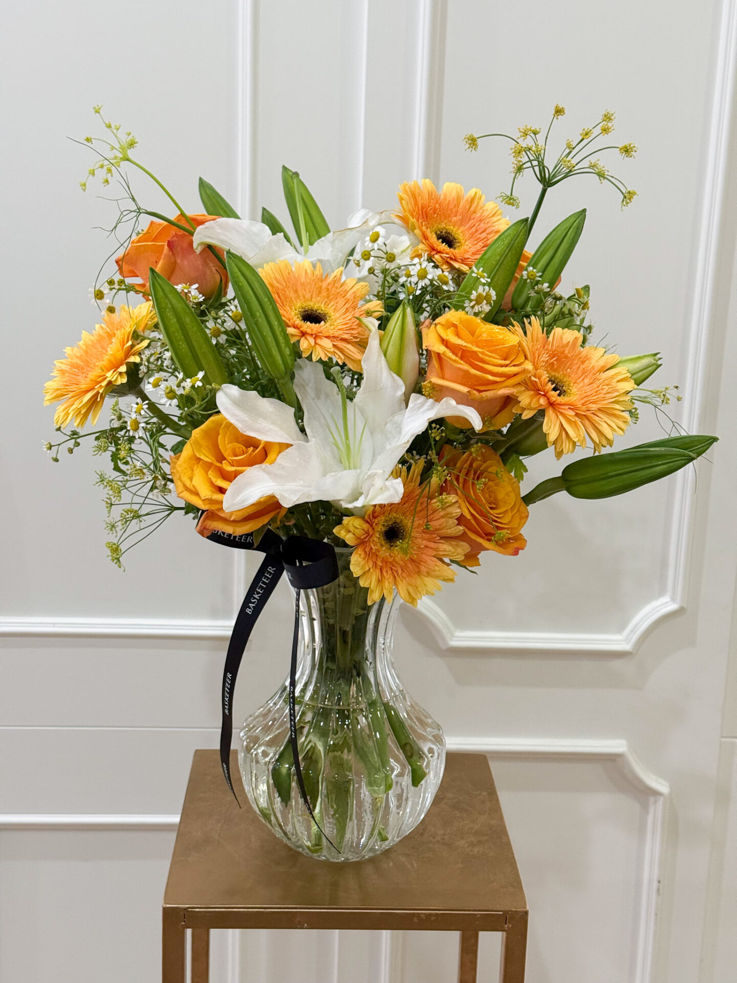 Bright vase with orange roses, orange gerberas, and white lilies in a clear glass vase, arranged with seasonal fillers.