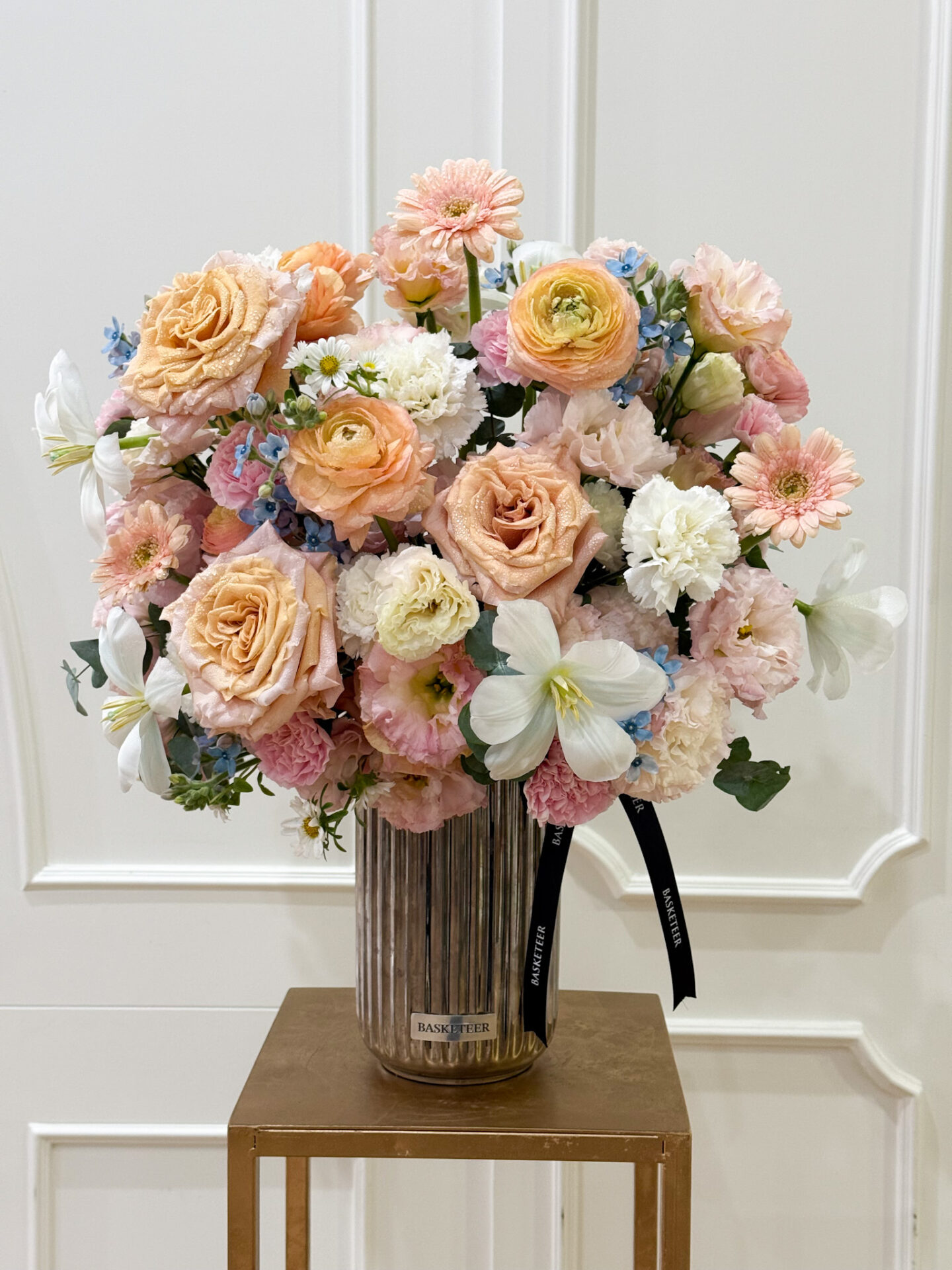 Luxury vase with peach roses, ranunculus, white lilies, pink carnations, and blue accent flowers in a ribbed glass vase.