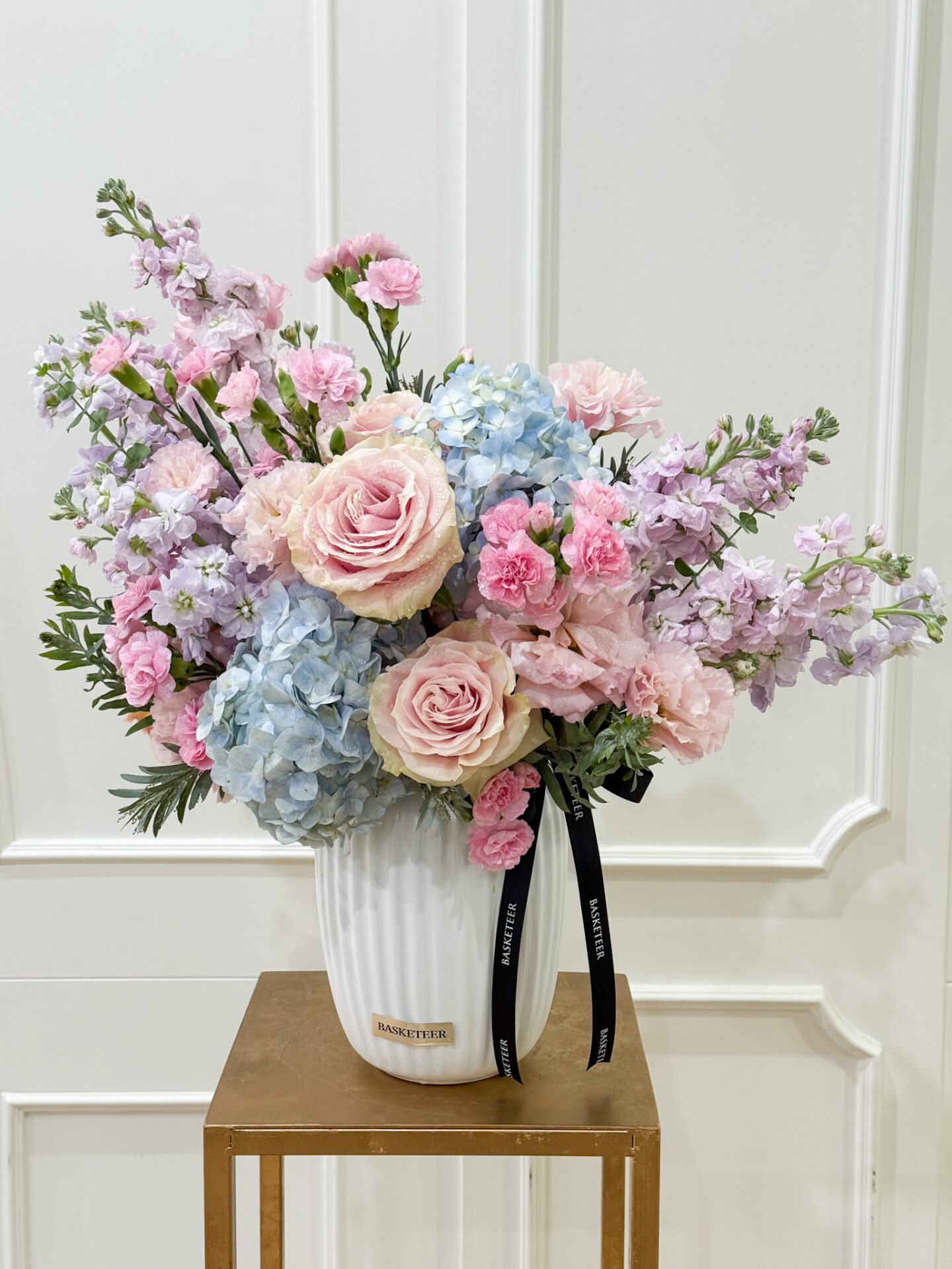 Elegant vase with pink roses, blue hydrangeas, pink carnations, and pastel stock flowers in a white ceramic vase.