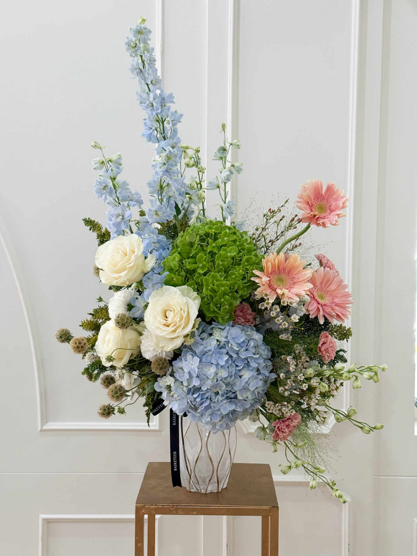 Luxury vase with blue delphinium, hydrangeas, white roses, pink gerberas, and greenery in pastel tones.