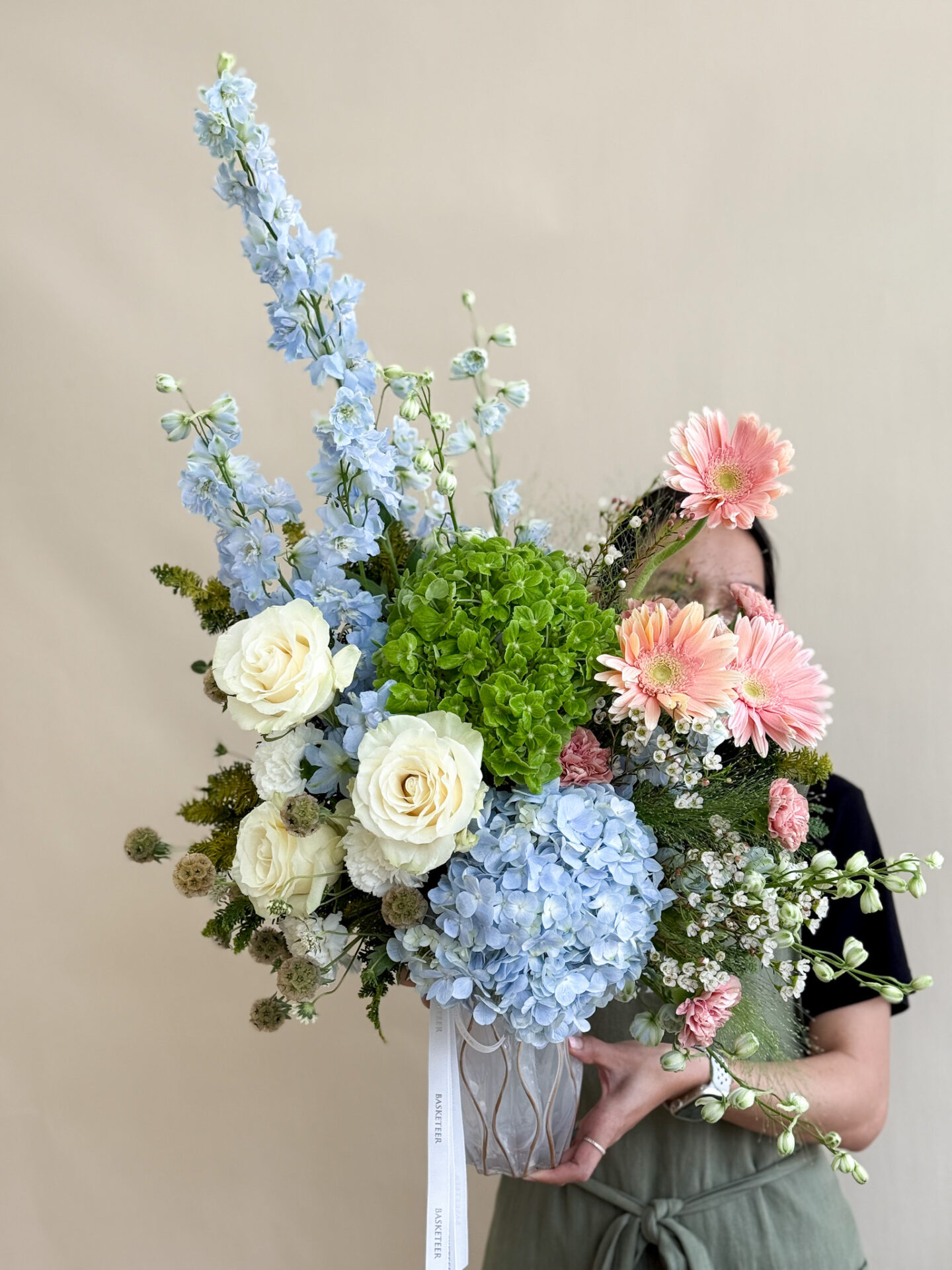 Tall luxury vase with blue delphinium, green hydrangeas, white roses, pink gerberas, and assorted blooms in pastel tones.