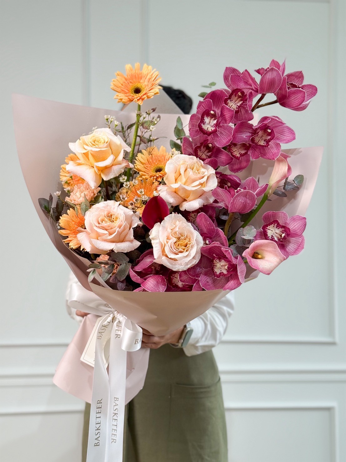 An exquisite Modern Luxe Bouquet of peach roses, orange gerberas, pink orchids, and greenery in pale pink wrap—Bangkok flower delivery.