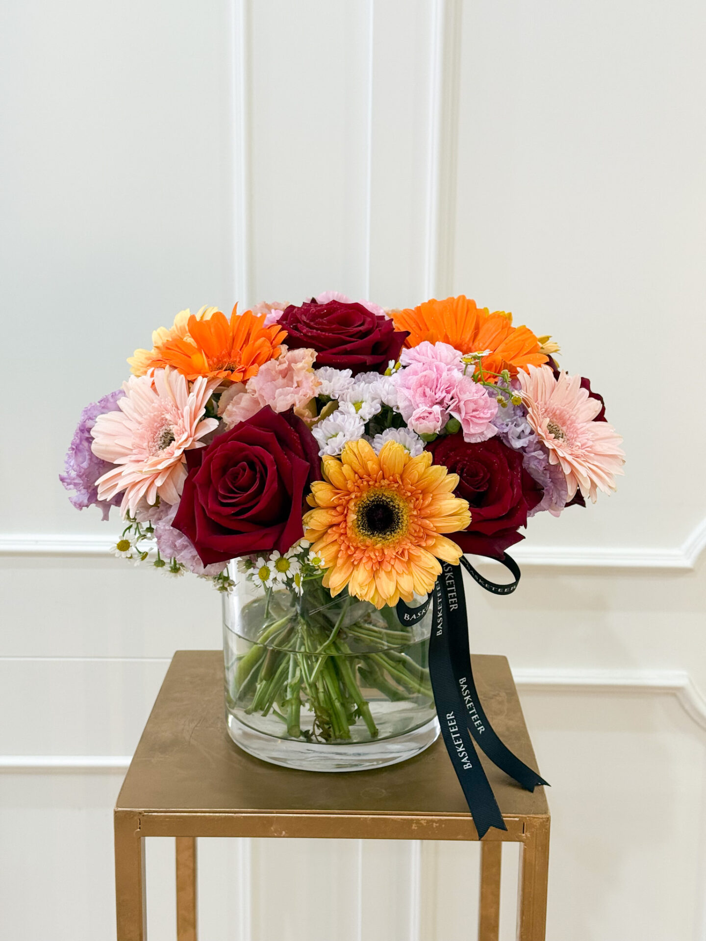 Glass vase with crimson red roses, orange and peach gerberas, pink carnations, purple statice, and chamomile flowers.