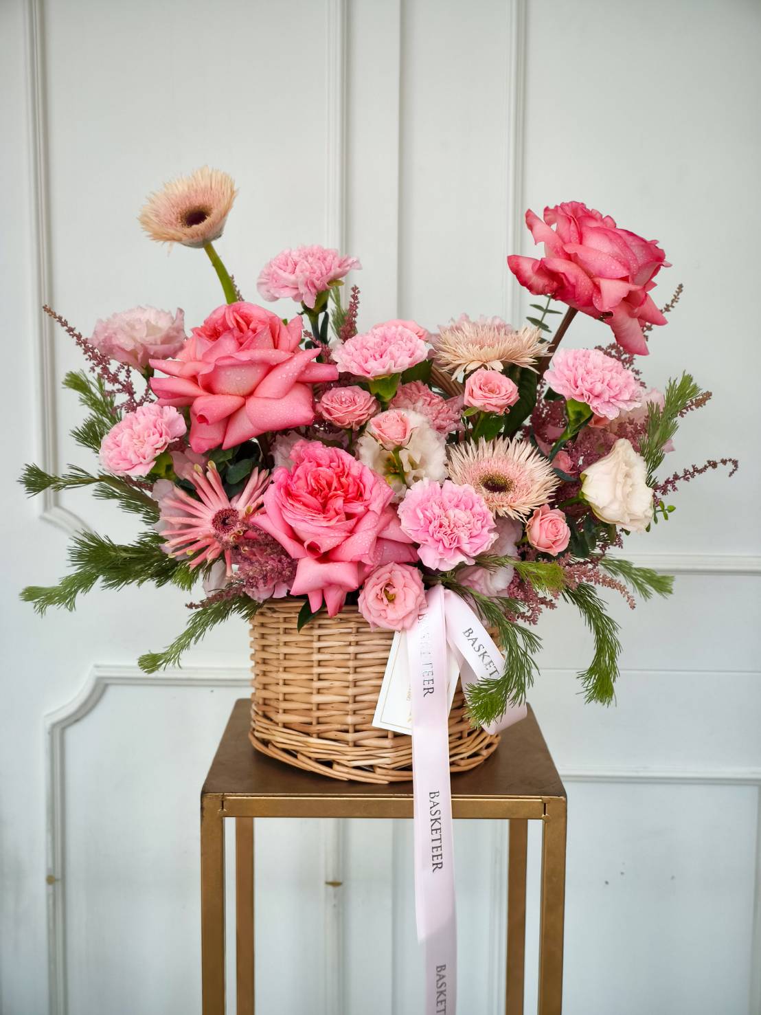 Woven basket with pink roses, carnations, gerberas, spray roses, and greenery, tied with a pink ribbon.