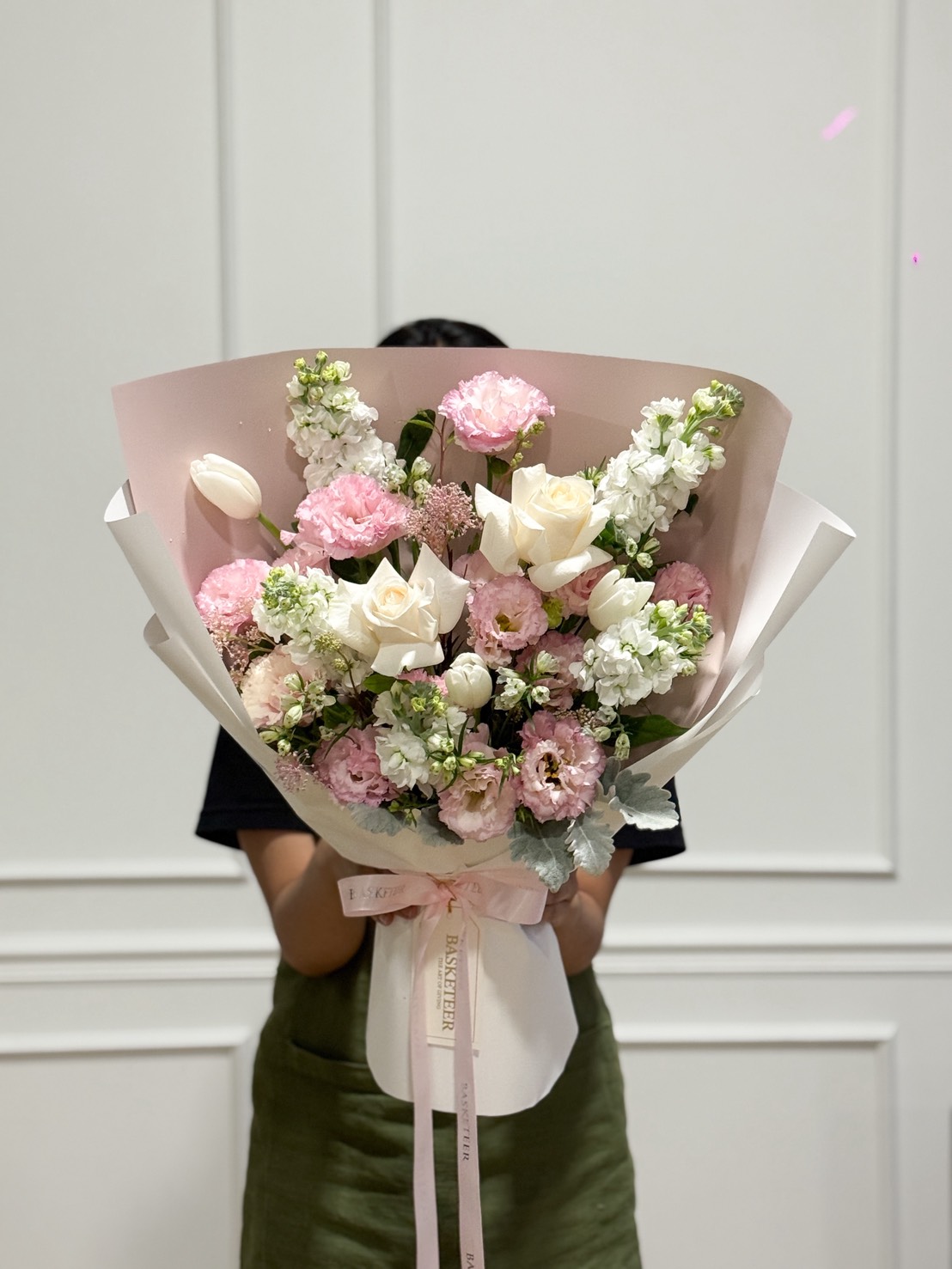 A hand presents a luxury bouquet of ivory and blush roses, white freesia, and carnations in pink wrap—Flowers by Basketeer, Bangkok.